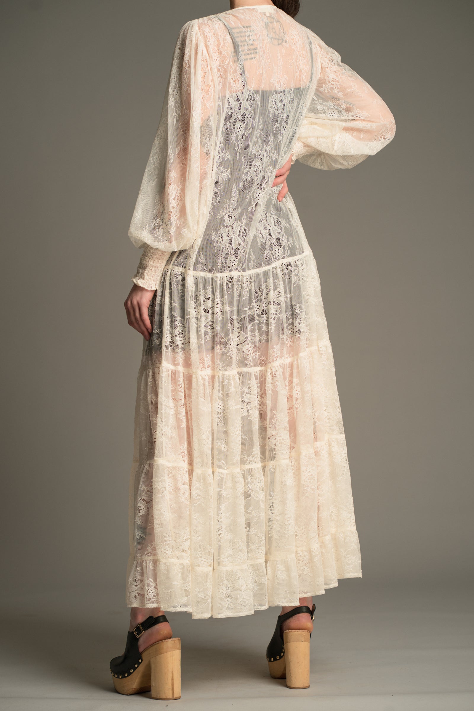 Back view of the Little Lies Rhiannon Ivory Maxi Dress in a studio setting, featuring sheer ivory lace fabric, long voluminous sleeves, and a tiered maxi‑length skirt.
