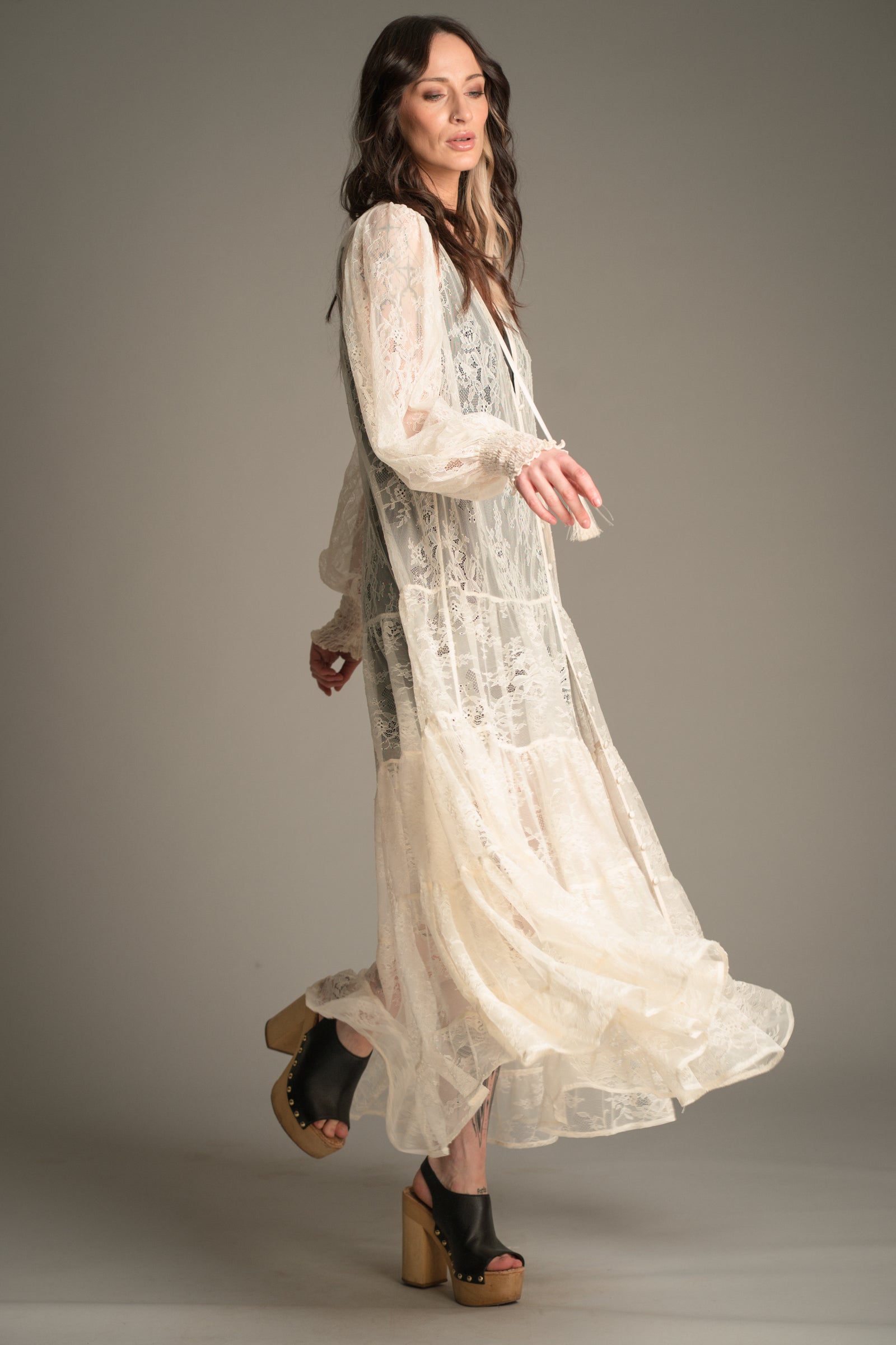 Side view of Victoria wearing the Little Lies Rhiannon Ivory Maxi Dress in sheer ivory lace, featuring long voluminous sleeves, tassel ties, and a flowing tiered skirt in motion, styled with wooden platform clogs.