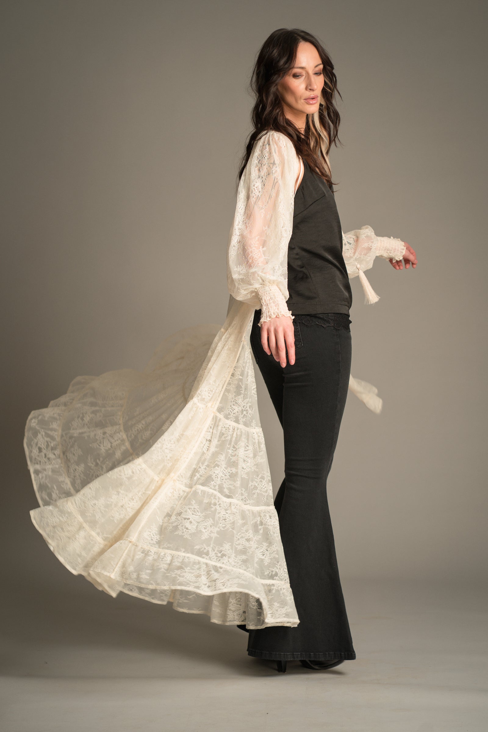 Side view of Victoria wearing the Little Lies Rhiannon Ivory Maxi Dress with flowing lace sleeves and a tiered lace skirt, worn open and styled over a black satin cami and denim flares.