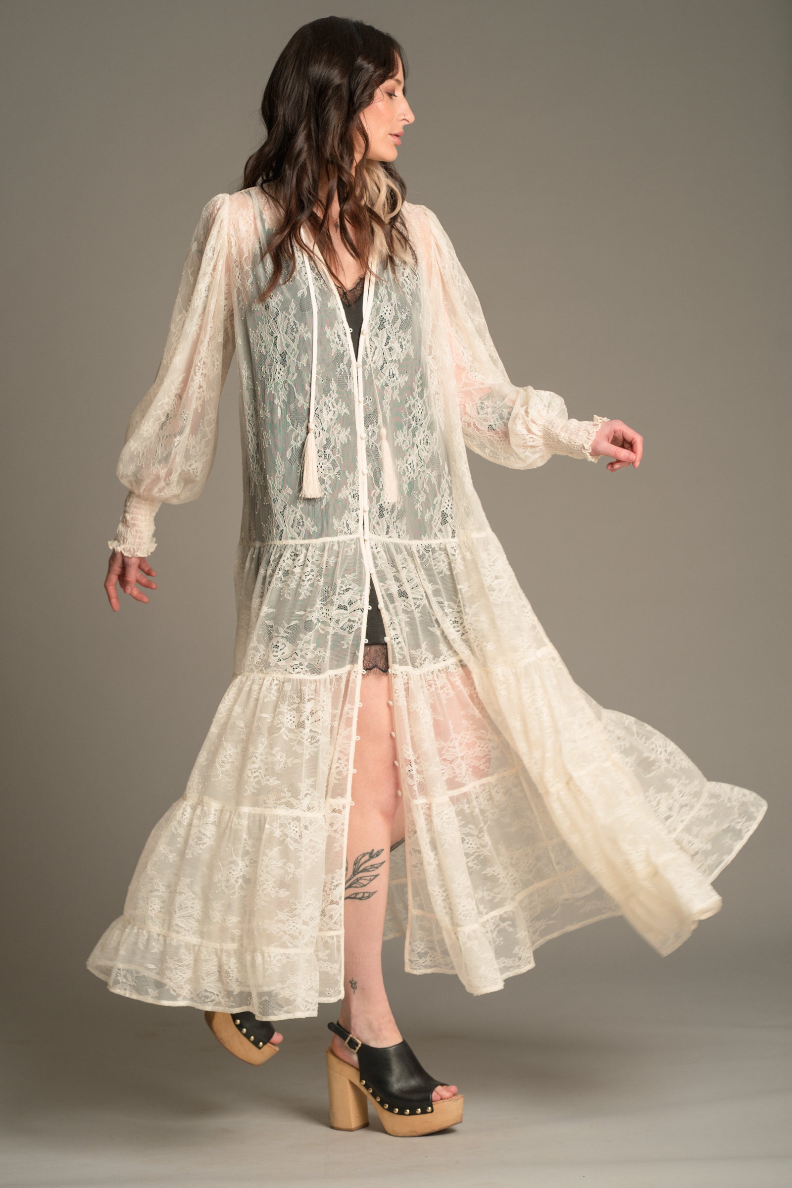 Front view of Victoria wearing the Little Lies Rhiannon Ivory Maxi Dress featuring sheer ivory lace fabric, long voluminous sleeves, tassel ties, and a tiered maxi‑length skirt worn over the Nightbird Satin Slip Dress as an underlayer.