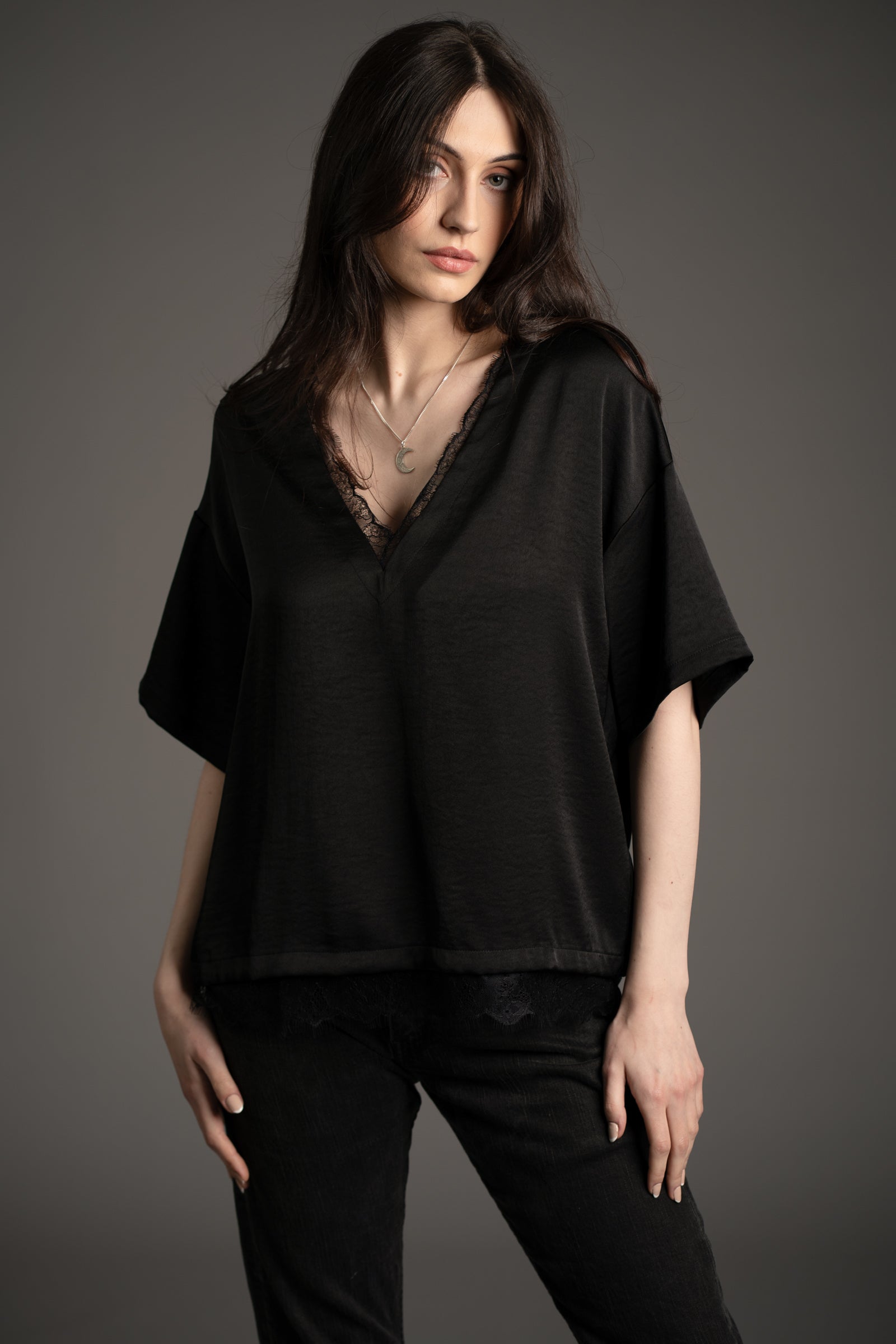Andie models Little Lies Roxy lace trim satin tee in black with deep V-neckline and delicate lace edging, styled with black jeans in studio setting