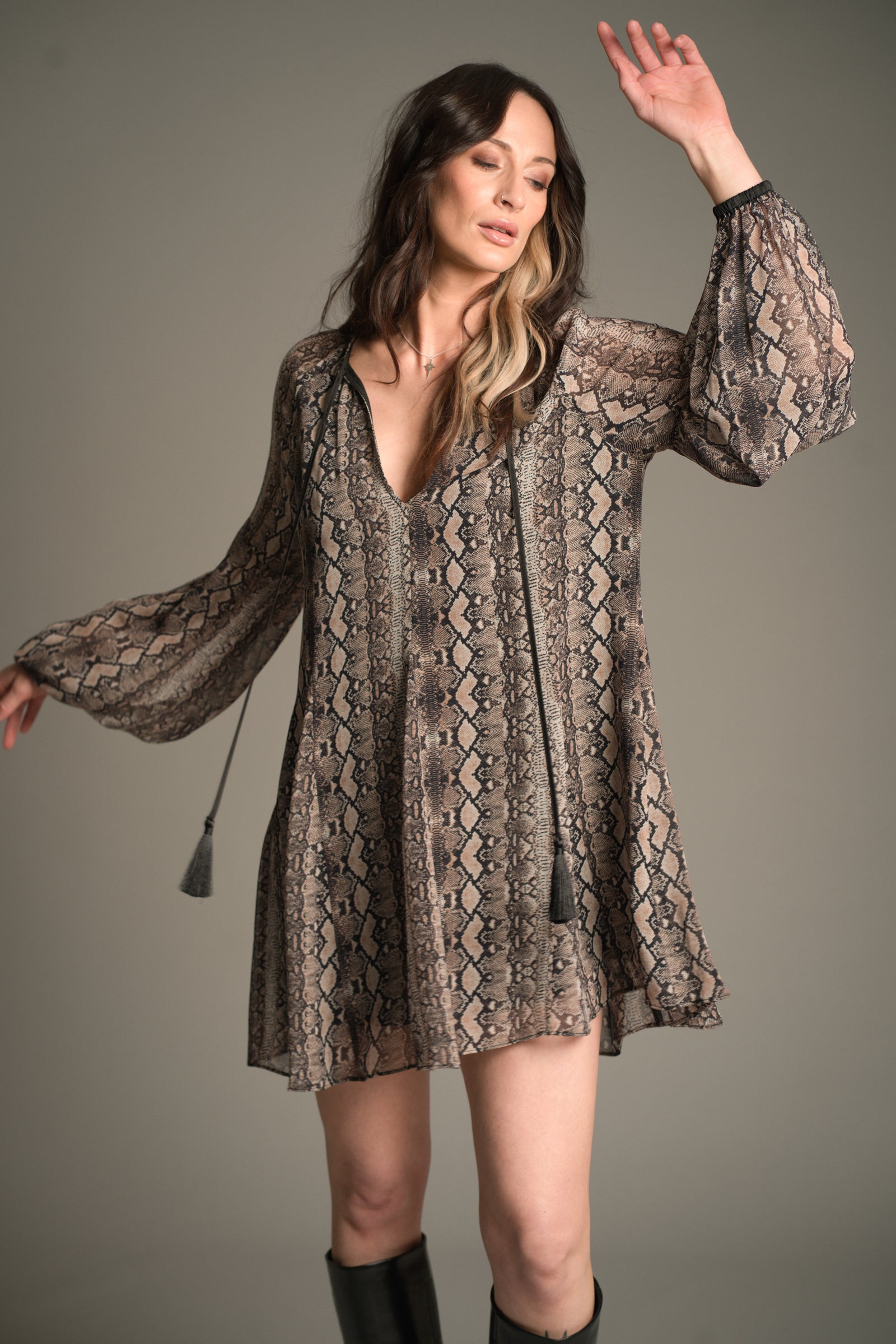 Front view of Victoria wearing the Little Lies Seeker Snake Mini Dress featuring a flowing snake‑print fabric, deep V‑neckline, long voluminous sleeves, and tassel ties, styled with knee‑high black boots.