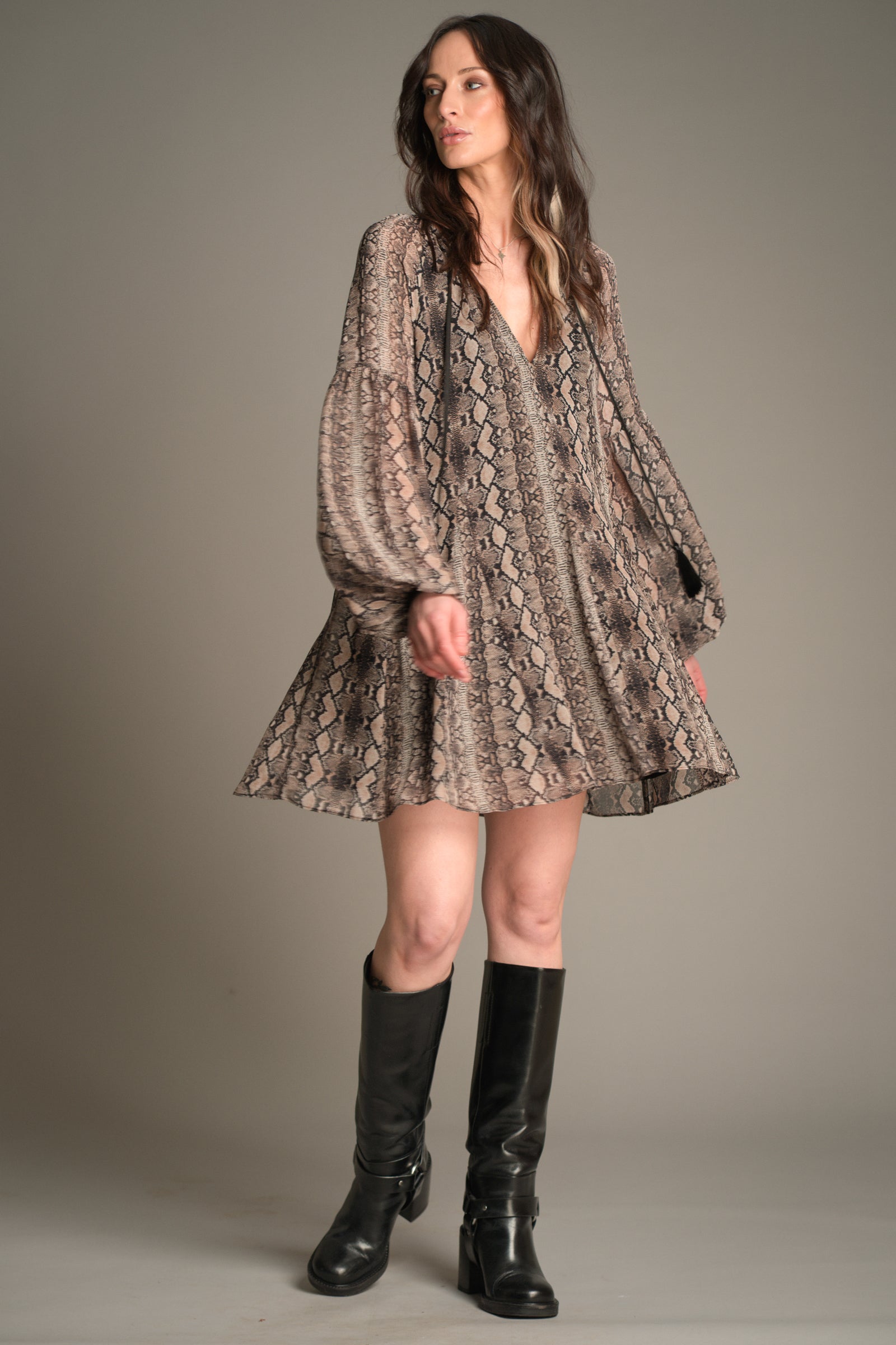 Front view of Victoria wearing the Little Lies Seeker Snake Mini Dress featuring a flowing snake‑print fabric, deep V‑neckline, and long voluminous sleeves, styled with knee‑high black boots.