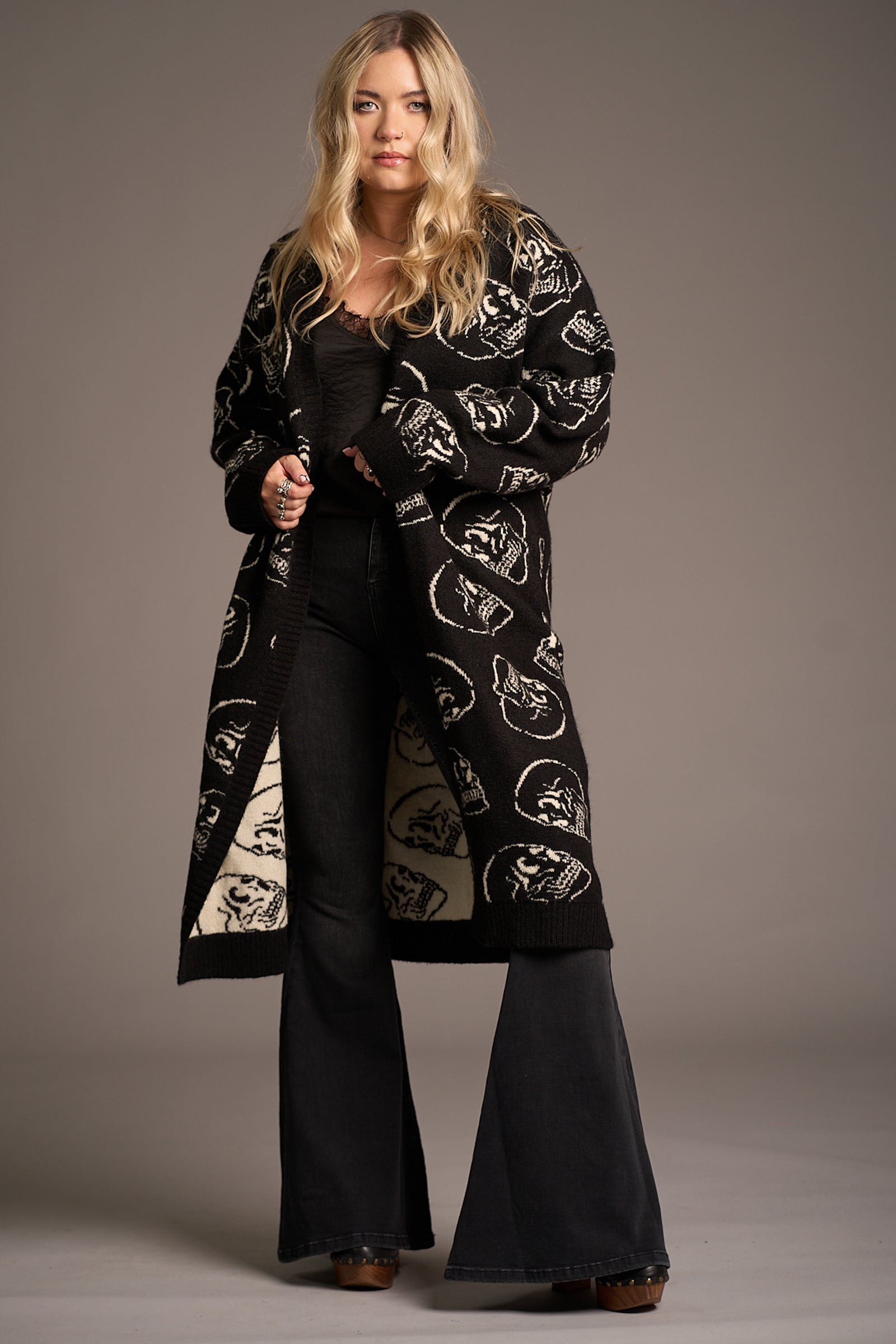 Erin wears the Little Lies Skull Knit Cardigan – longline black knit with bold white skull motifs, styled open over a black top and flared denim for a layered, edgy studio look.