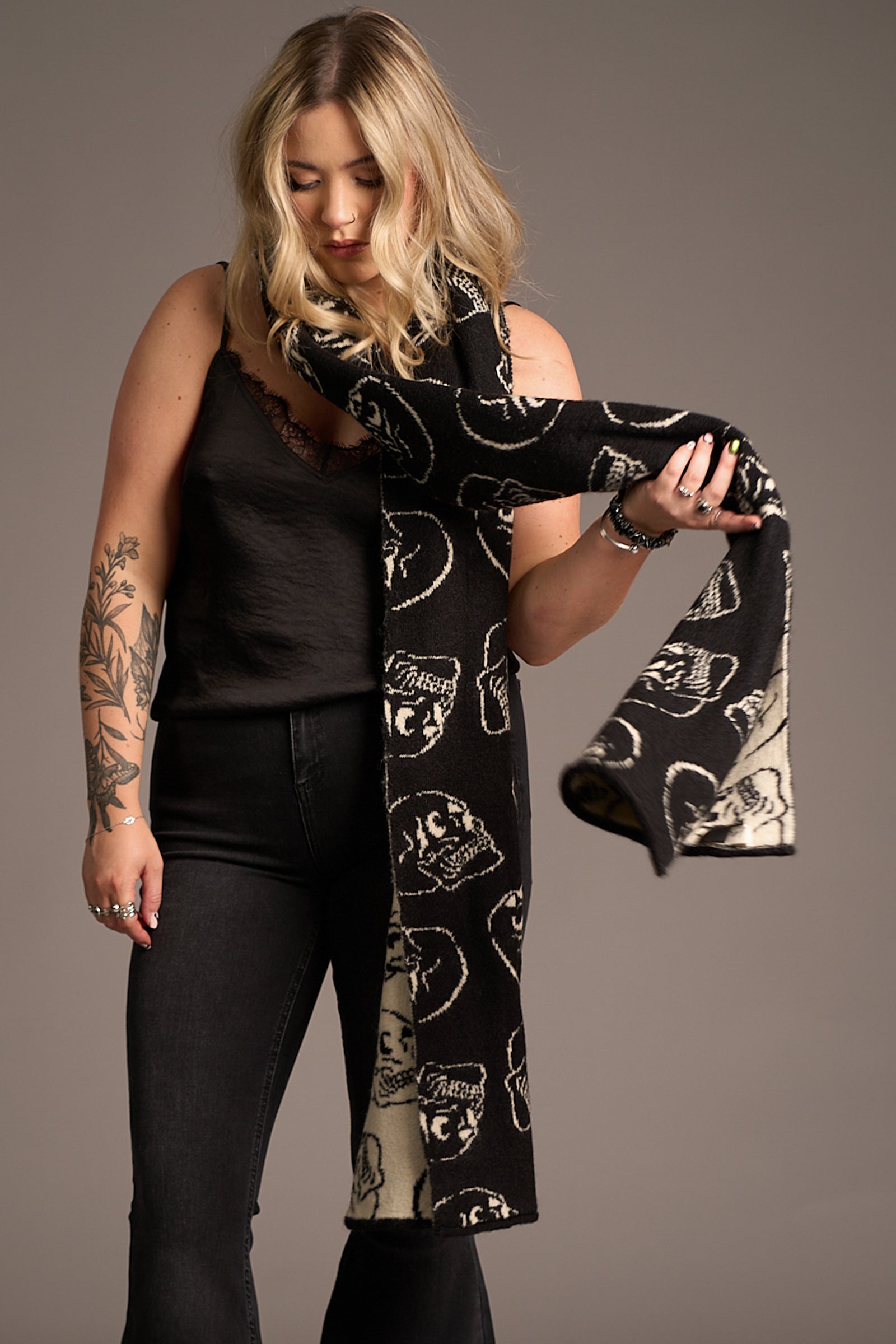 Erin wears Little Lies Skull Knit Scarf – long black knitted scarf with bold white skull motif, styled with an a black lace cami and denim flares in a minimalist studio setting for a statement winter look.