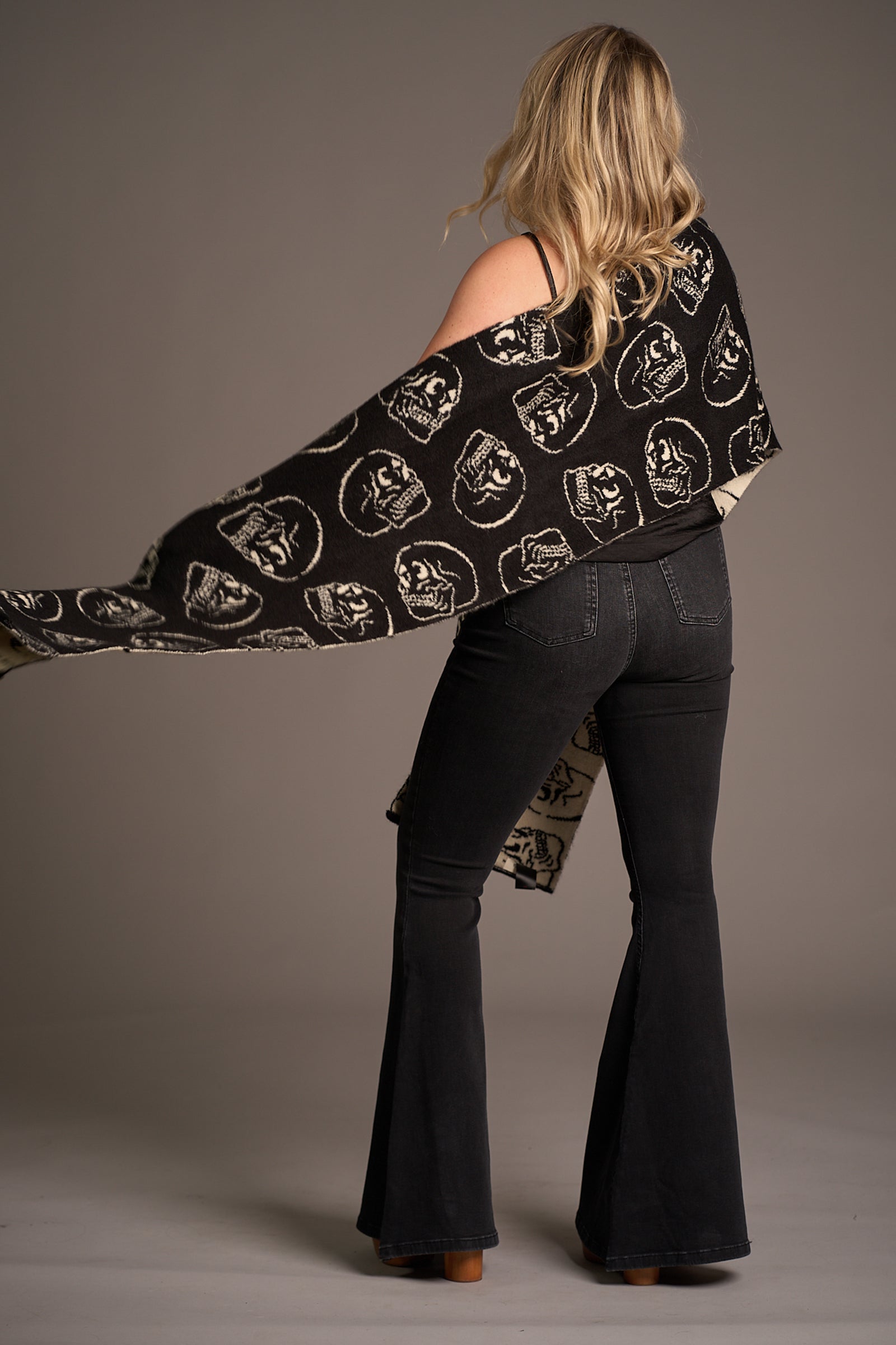 Back view of Erin wearing Little Lies Skull Knit Scarf – black shawl-style knit with bold white skull motifs, draped over shoulders and paired with flared denim for a layered studio look.