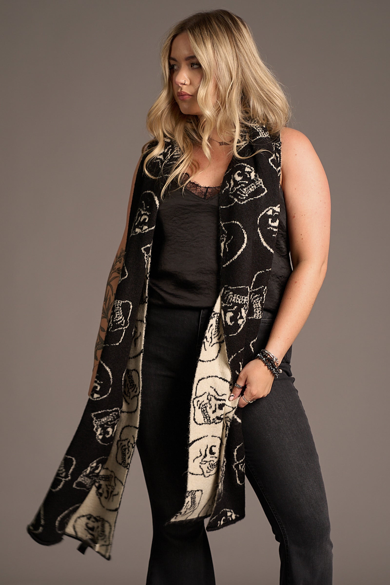Front view of Little Lies Skull Knit Scarf – black knit with bold white skulls and a contrasting light reverse side, styled draped over shoulders to reveal dual-tone detail in a studio setting.