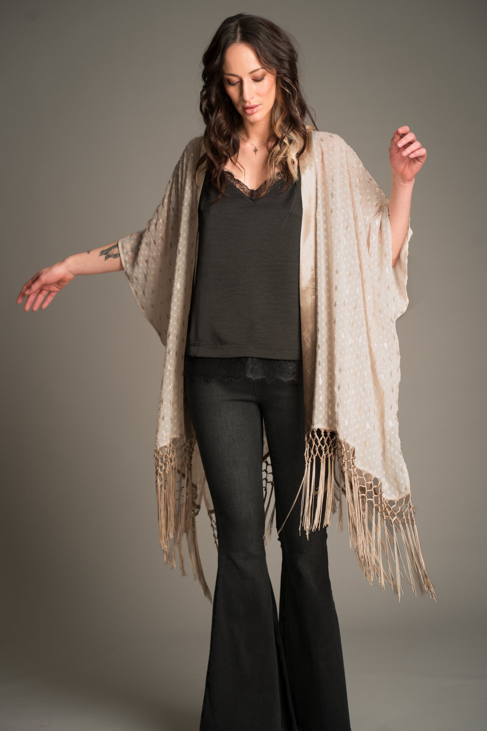 Studio shot of Victoria wearing the Little Lies Songbird Ivory Kimono featuring a soft ivory draped fabric with subtle textured detailing and long fringe trim, styled over a black satin cami and flared denim trousers