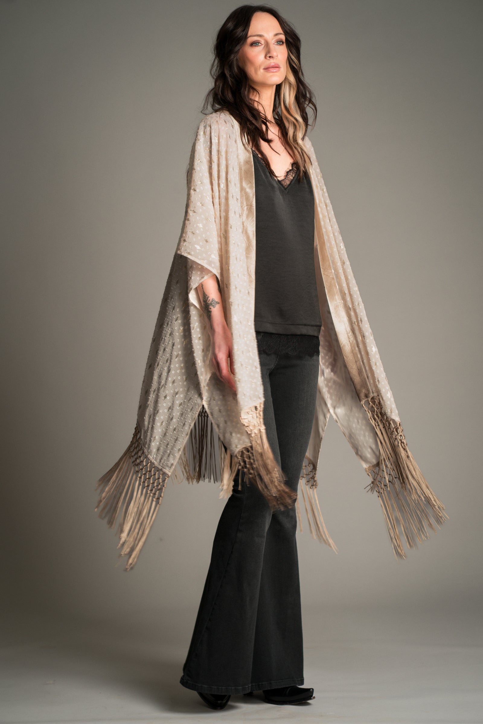 Front view of Victoria wearing the Little Lies Songbird Ivory Kimono in a studio setting, featuring sheer ivory textured fabric with metallic detailing and long knotted fringe trim, styled over a black satin cami and denim flares.