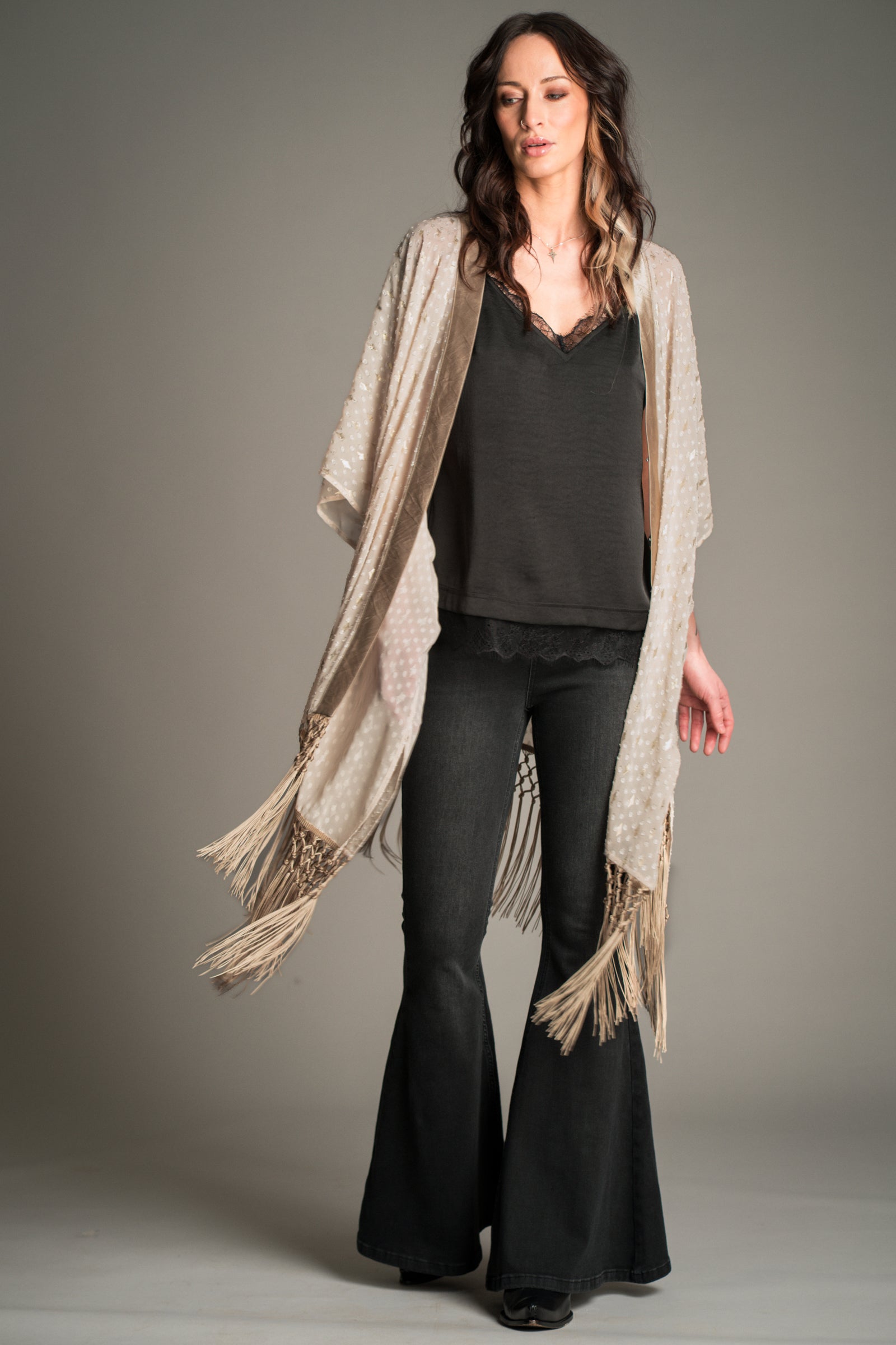 Studio shot of Victoria wearing the Little Lies Songbird Ivory Kimono featuring a soft ivory draped fabric with subtle textured detailing and long fringe trim, styled over a black satin cami and flared denim trousers