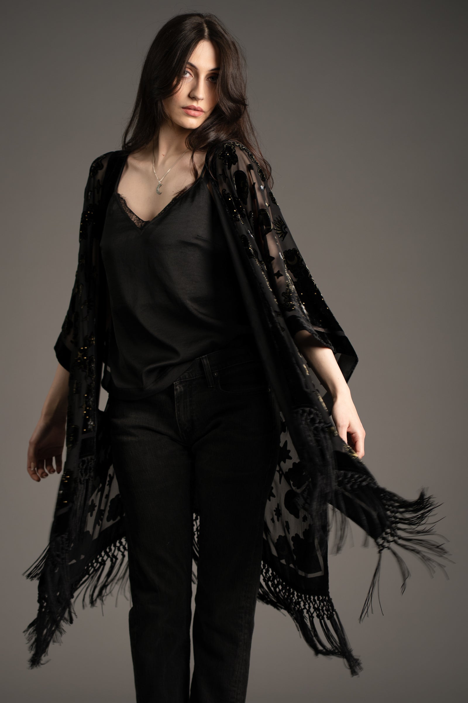Andie models Little Lies Suzi Skull devore kimono in black with skull and celestial pattern, sheer fabric and fringe hem, styled over black camisole and dark jeans in studio setting