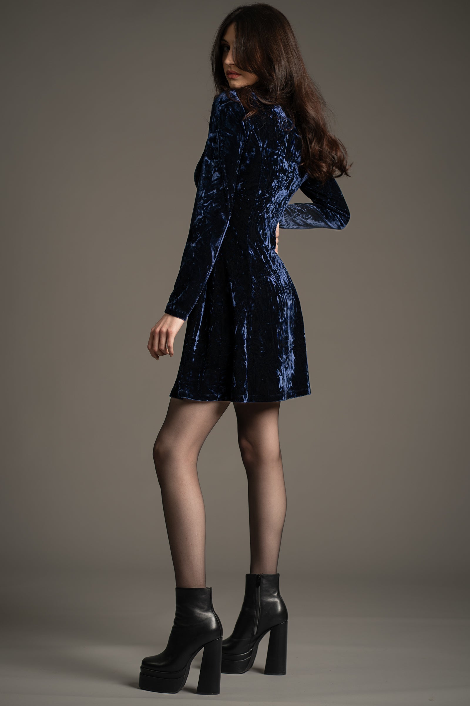Side view of Andie modelling Little Lies Sweet Jane Midnight Crushed Mini Dress in dark blue velvet, long sleeves, styled with black platform boots, studio setting