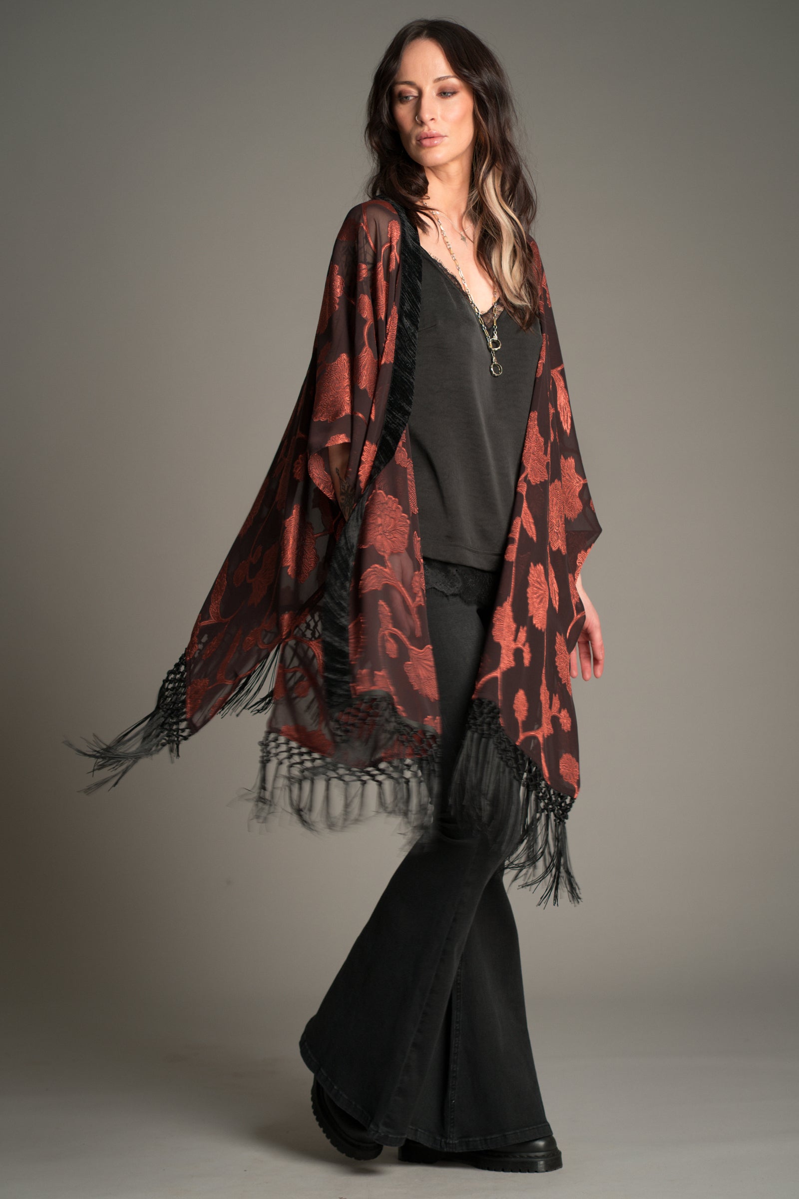 Victoria wears Little Lies Venus Fringed Kimono in black and rust floral patterned fabric with long fringe detailing, styled over a black cami and flared trousers.