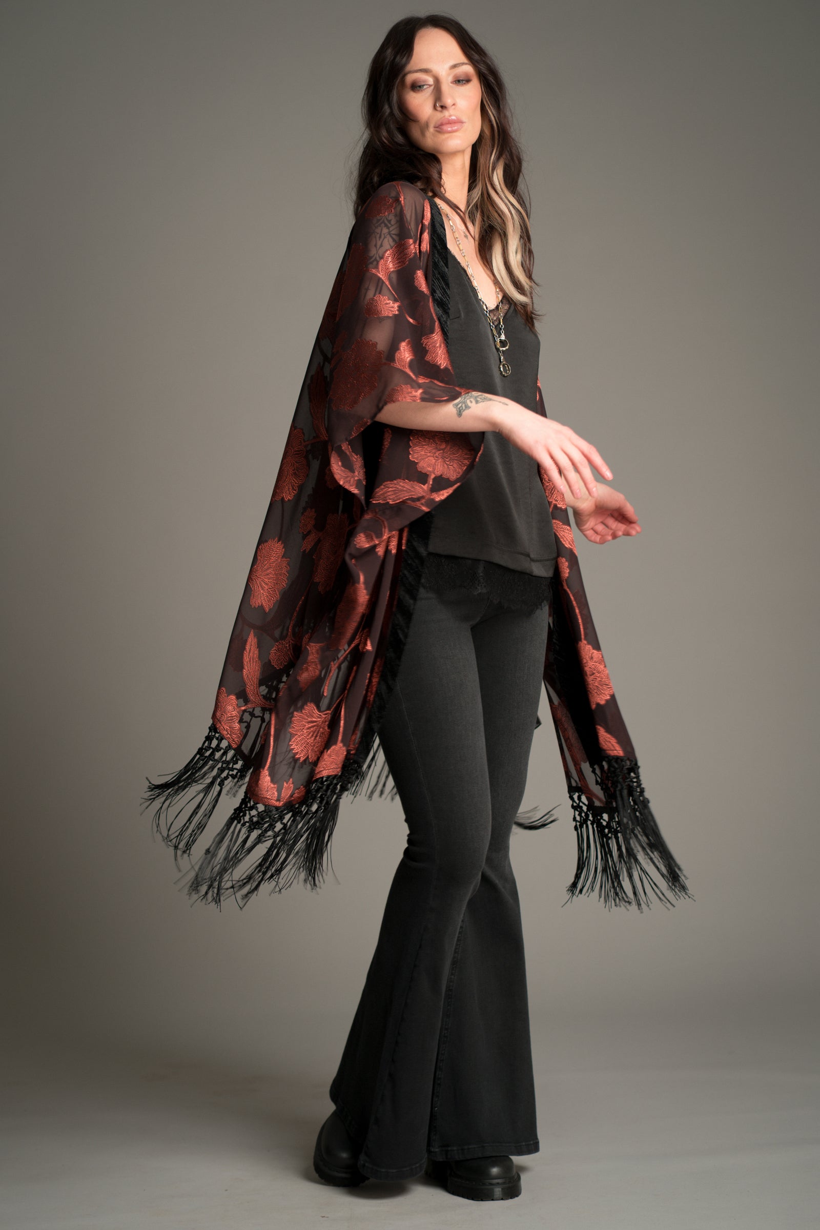 Victoria wears Little Lies Venus Fringed Kimono in black and rust floral patterned fabric with long fringe detailing, styled over a black cami and flared trousers.