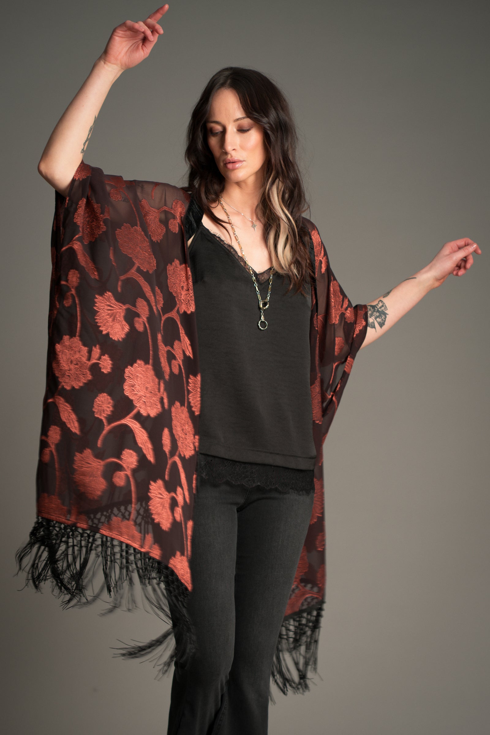 Victoria wears Little Lies Venus Fringed Kimono in black and rust floral patterned fabric with long fringe detailing, styled over a black cami and flared trousers.