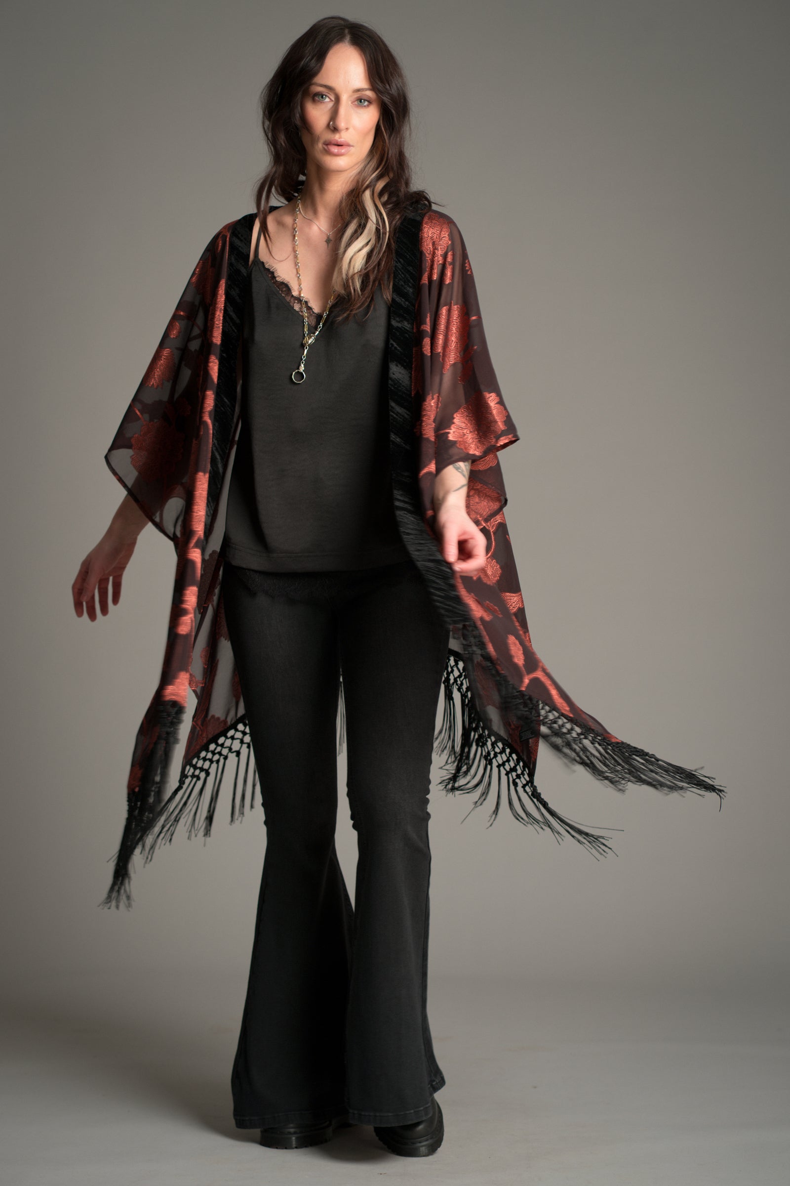 Victoria wears Little Lies Venus Fringed Kimono in black and rust floral patterned fabric with long fringe detailing, styled over a black cami and flared trousers.