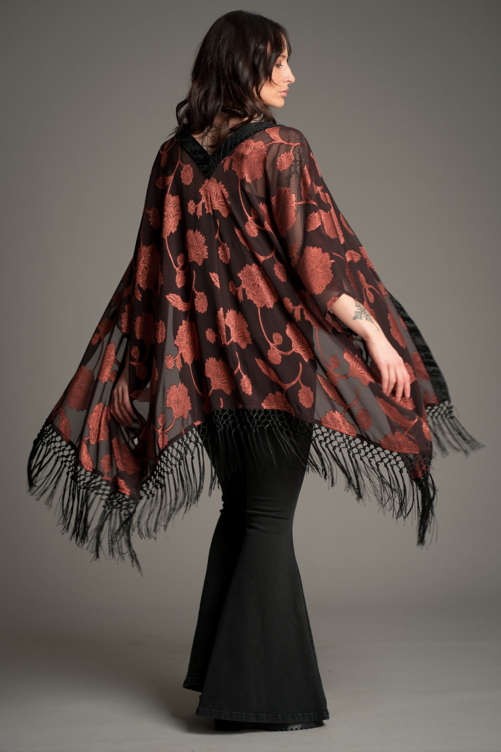 Back view of the Little Lies Venus Fringed Kimono in black with rust floral pattern and long black fringe detailing, styled with flared denim trousers.