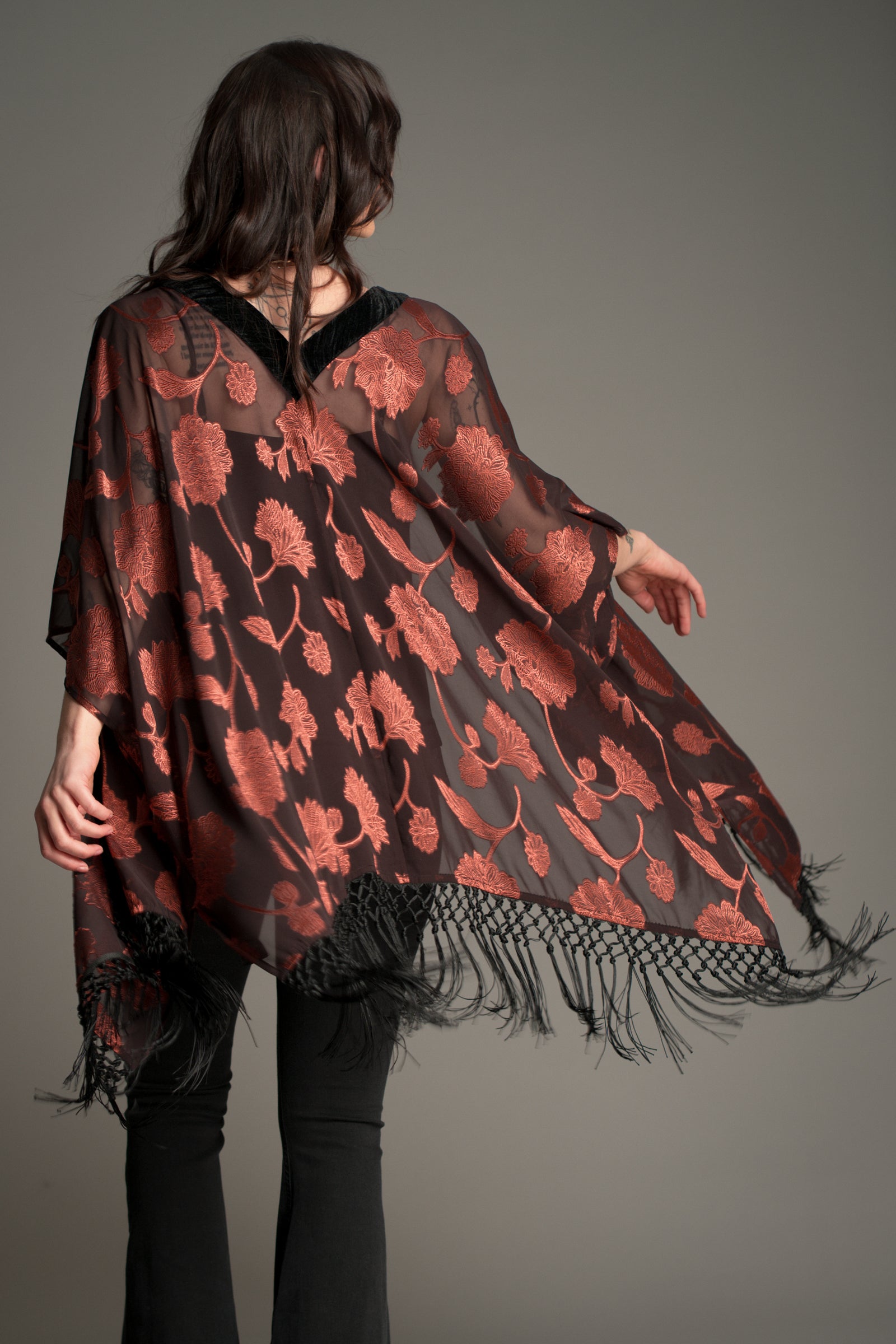 Back view of the Little Lies Venus Fringed Kimono in black with rust floral pattern and long black fringe detailing, styled with flared denim trousers.