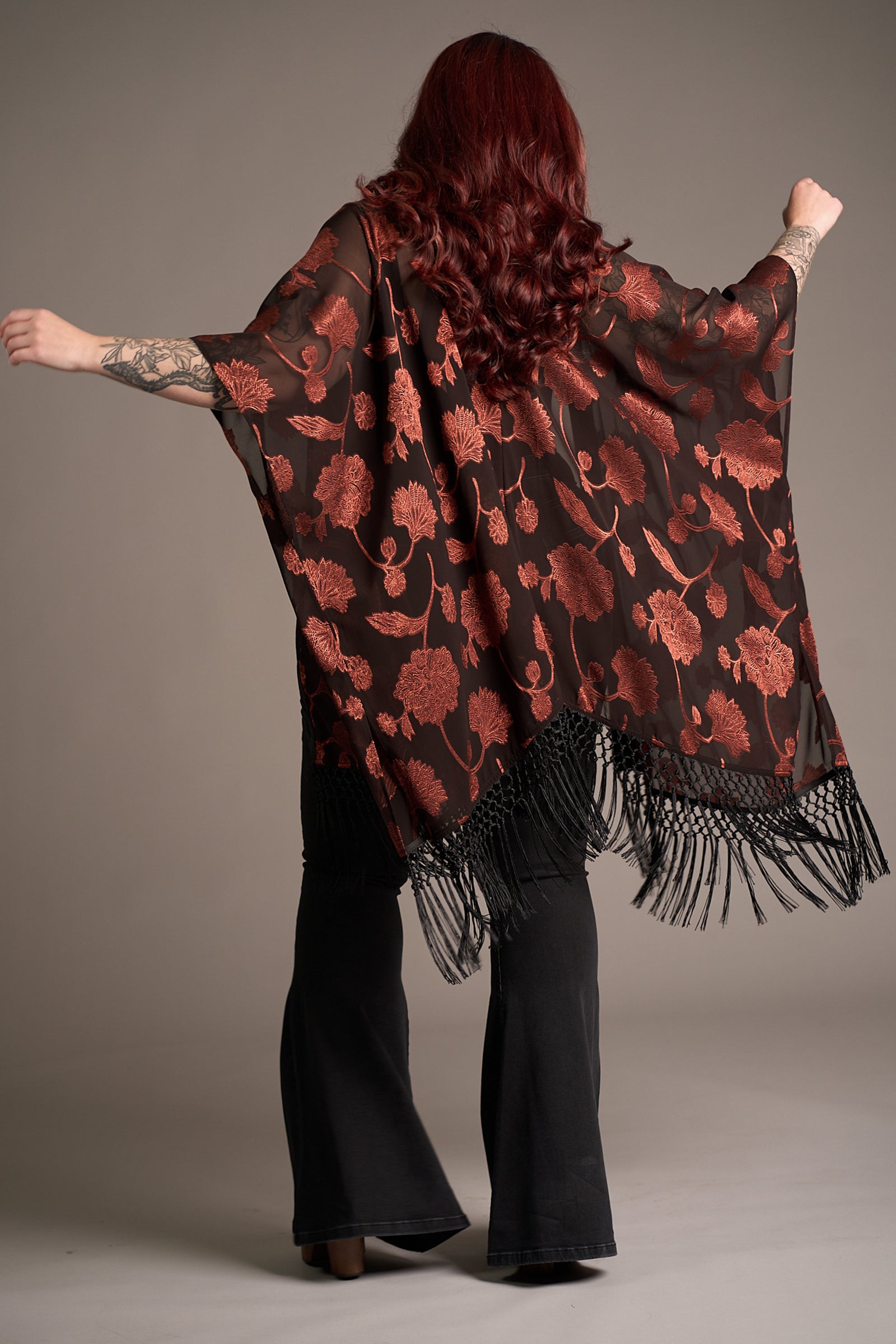 Taylor showing the back of the Venus Fringed Kimono with black sheer fabric, red floral print, and fringe hem. Stevie Nicks inspired styling studio