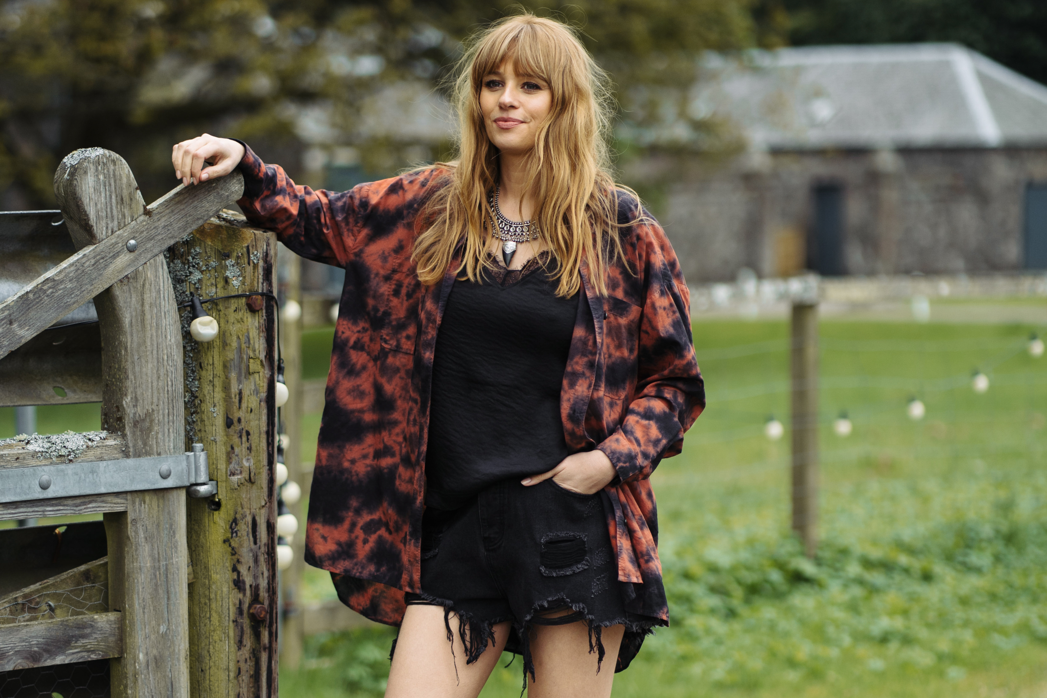 Model in Zephyr red and black tie-dye shirt over black crop top and shorts, leaning on wooden fence, festival style.
