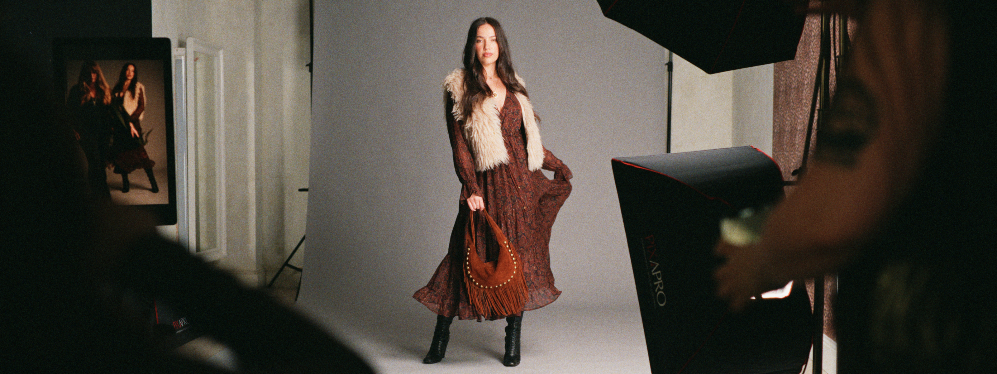 Behind the scenes of the Little Lies autumn campaign featuring the Dark Paradise Paisley Maxi Dress styled with a faux fur gilet and suede fringe bag during a fashion photoshoot.