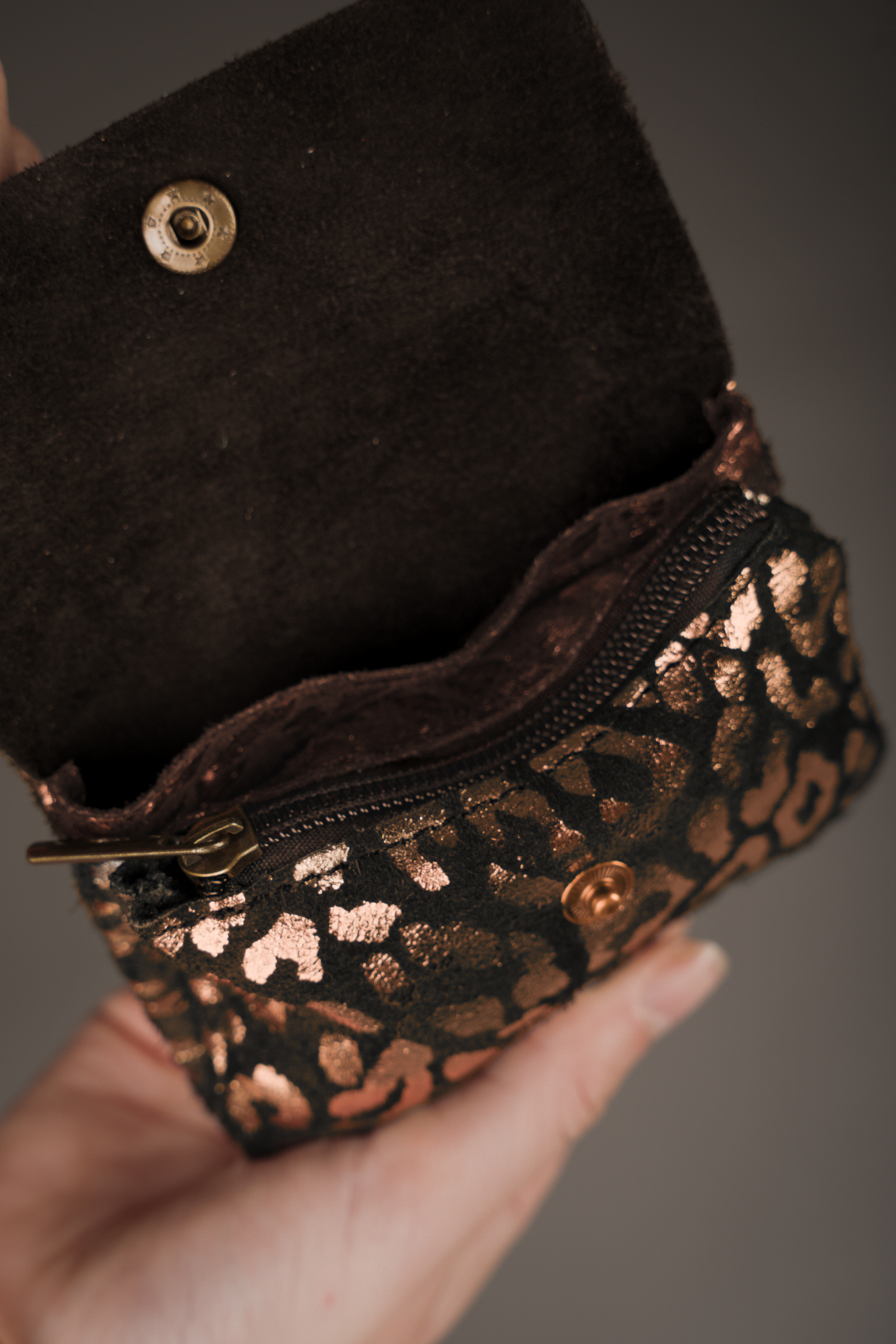 Open copper metallic suede purse with black animal‑print texture, showing zip compartment and button flap, held against a grey background.
