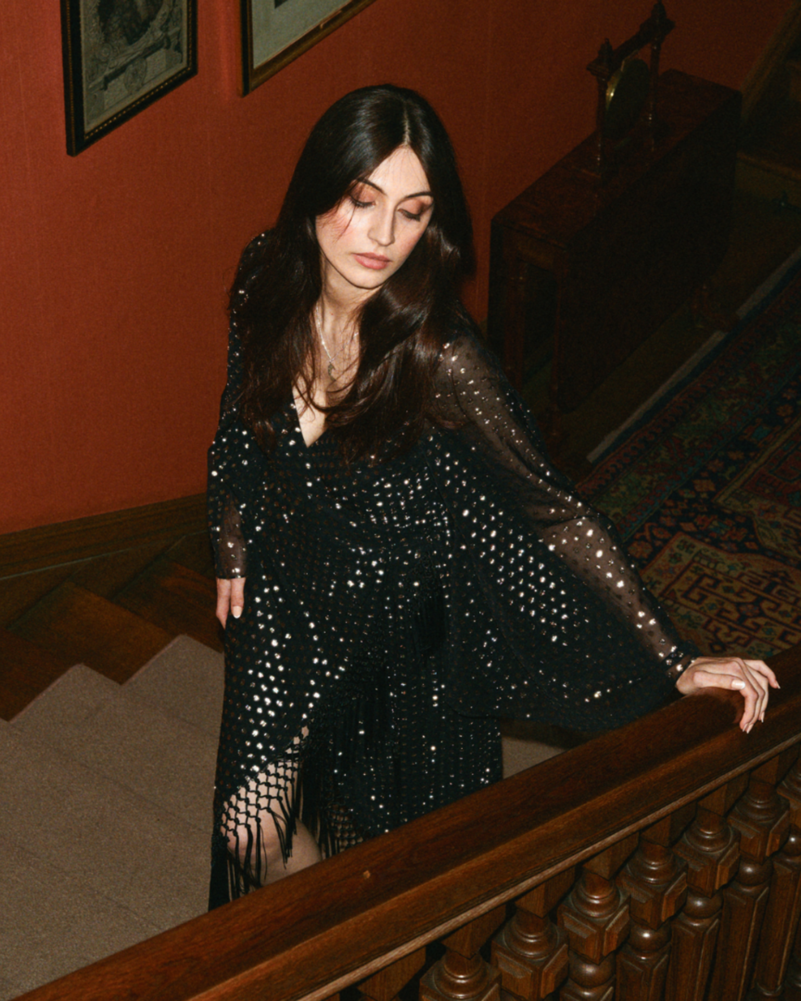 Woman in a black sequined dress standing on a staircase with red walls.