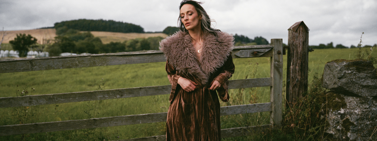 Model wearing the Little Lies Sienna Velvet Kimono Jacket in a Spellbound collection campaign shoot, featuring rich bronze velvet and a faux fur collar in a rural countryside setting.