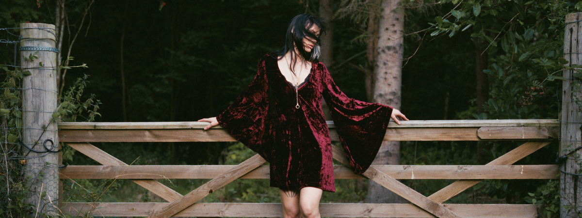 Model wearing the Little Lies Sara Merlot Velvet Mini Dress in a Spellbound collection campaign shoot, featuring luxurious crushed velvet and dramatic bell sleeves in a woodland setting.
