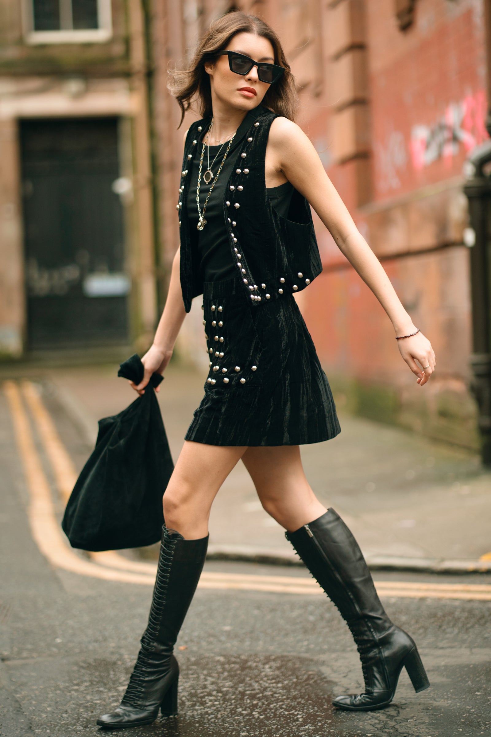 Model wearing Little Lies Lennon studded velvet mini skirt styled with black top, matching waistcoat and knee-high boots for edgy street-style outfit
