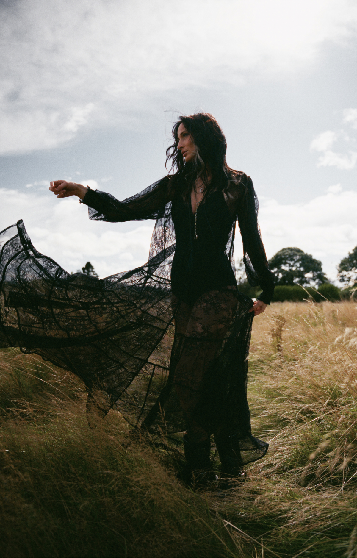 Model wearing the Little Lies Rhiannon Black Maxi Dress in a Spellbound collection campaign shoot, featuring sheer lace detailing and flowing black silhouette in a countryside setting.