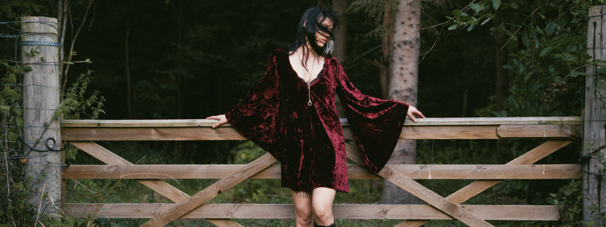 Model wearing the Little Lies Sara Merlot Velvet Mini Dress in a Spellbound collection campaign shoot, featuring luxurious crushed velvet and dramatic bell sleeves in a woodland setting.