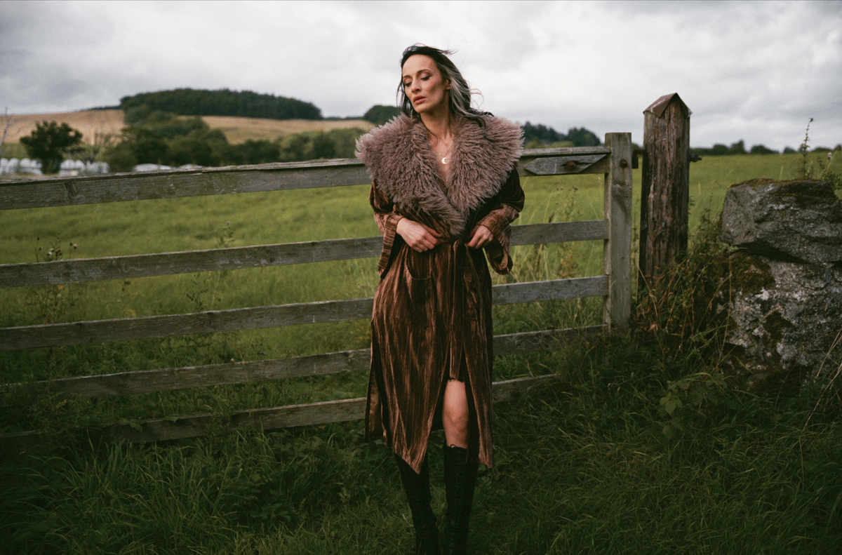 Model wearing the Little Lies Sienna Velvet Kimono Jacket in a Spellbound collection campaign shoot, featuring rich bronze velvet and a faux fur collar in a rural countryside setting.
