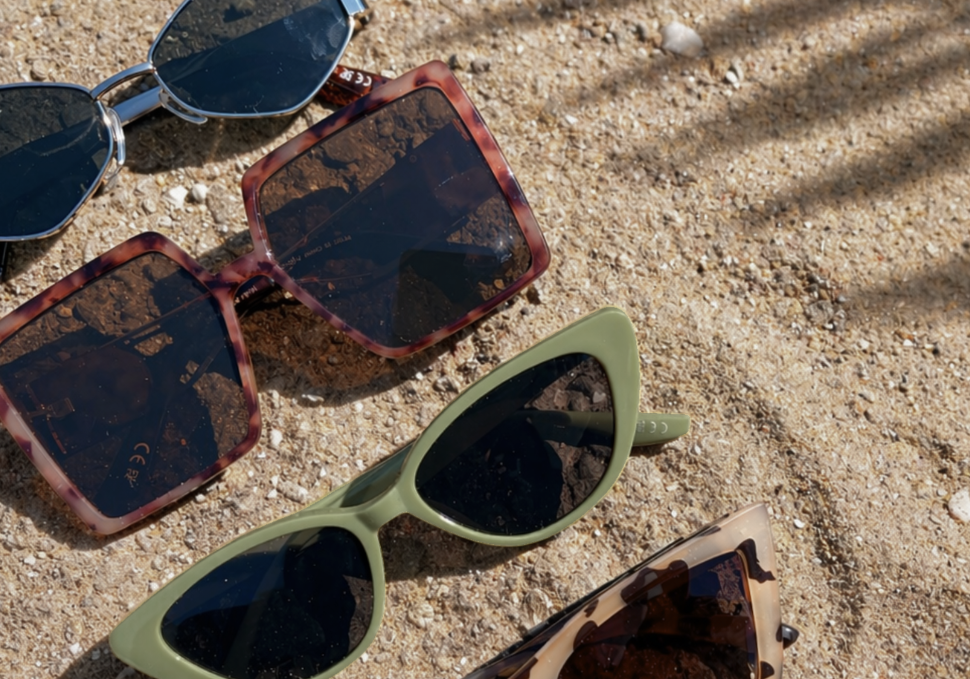 Little Lies sunglasses collection flat lay on sand featuring an assortment of boho summer styles beach.
