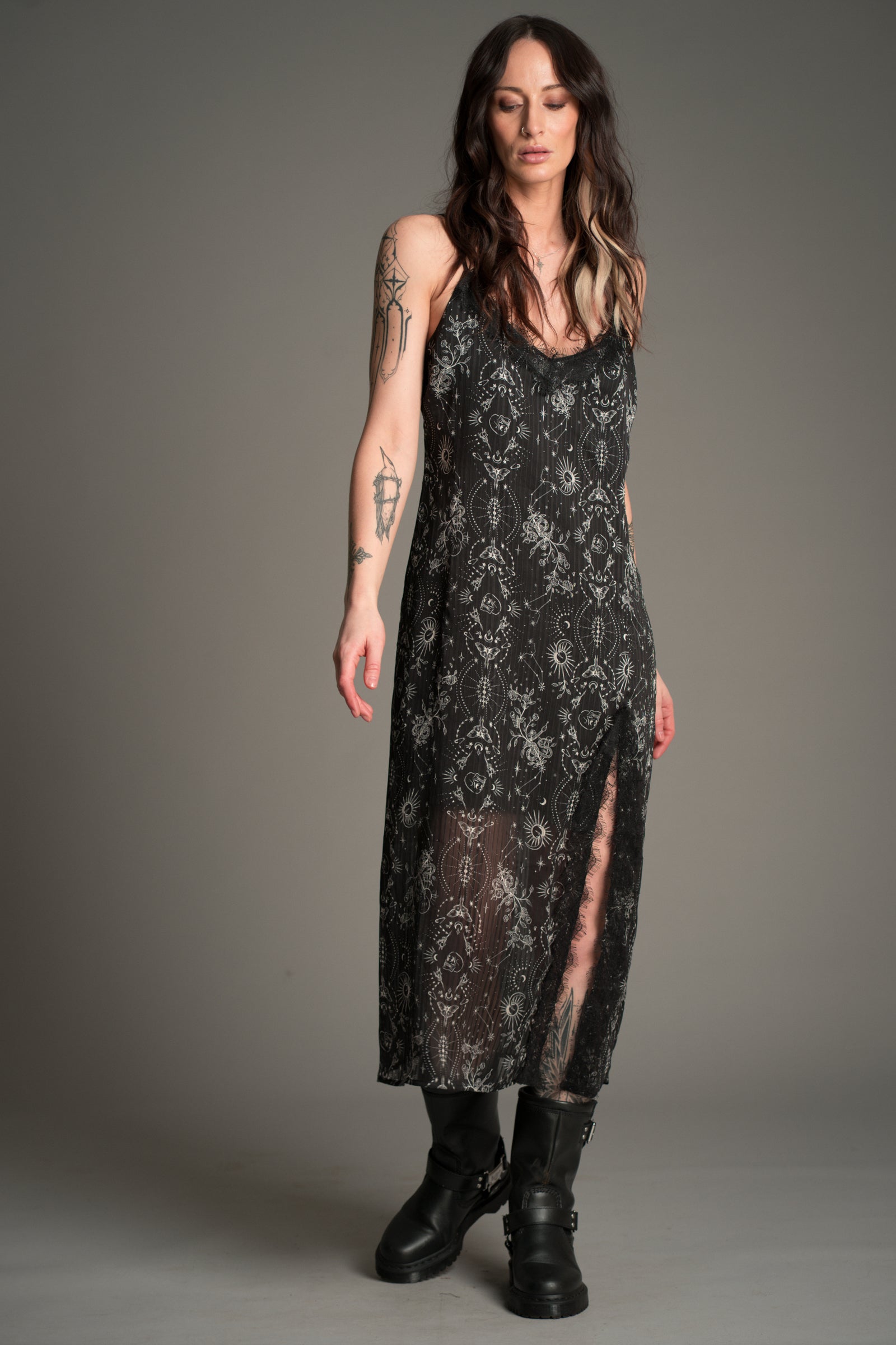 Front view of Victoria in the Little Lies Tattoo Slip Dress in black patterned lace with sheer detailing and a side split, styled with chunky black boots.