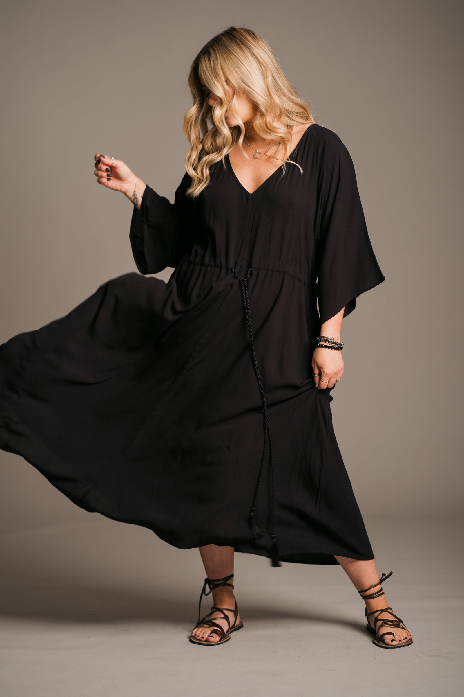 Model Erin wearing the Little Lies Blackbird Tie Waist Midi Dress, featuring a flattering V-neckline, batwing sleeves, an adjustable tie waist, and a flowing midi-length kaftan silhouette. This lightweight, drapey dress is styled with black ankle-tie sandals, exuding effortless bohemian elegance against a neutral backdrop.