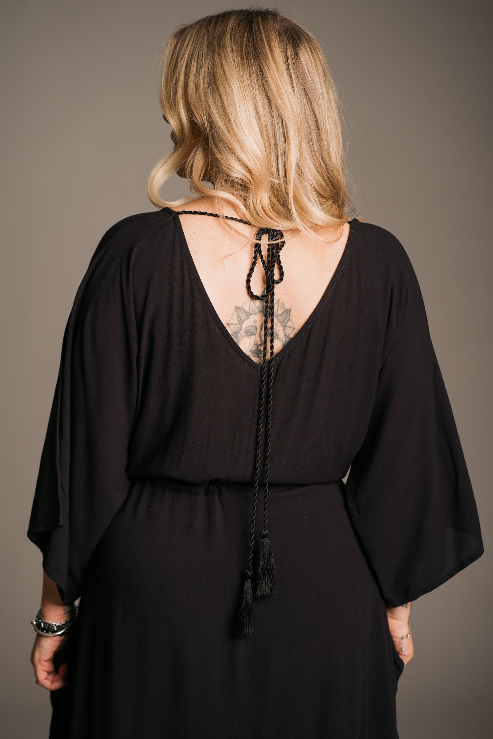 Model Erin wearing the Little Lies Blackbird Tie Waist Midi Dress showing the back of the dress, featuring a flattering V-neckline on the back a tie cord across the upper back, batwing sleeves, an adjustable tie waist, and a flowing midi-length kaftan silhouette. This lightweight, drapey dress is styled with black ankle-tie sandals, exuding effortless bohemian elegance against a neutral backdrop.