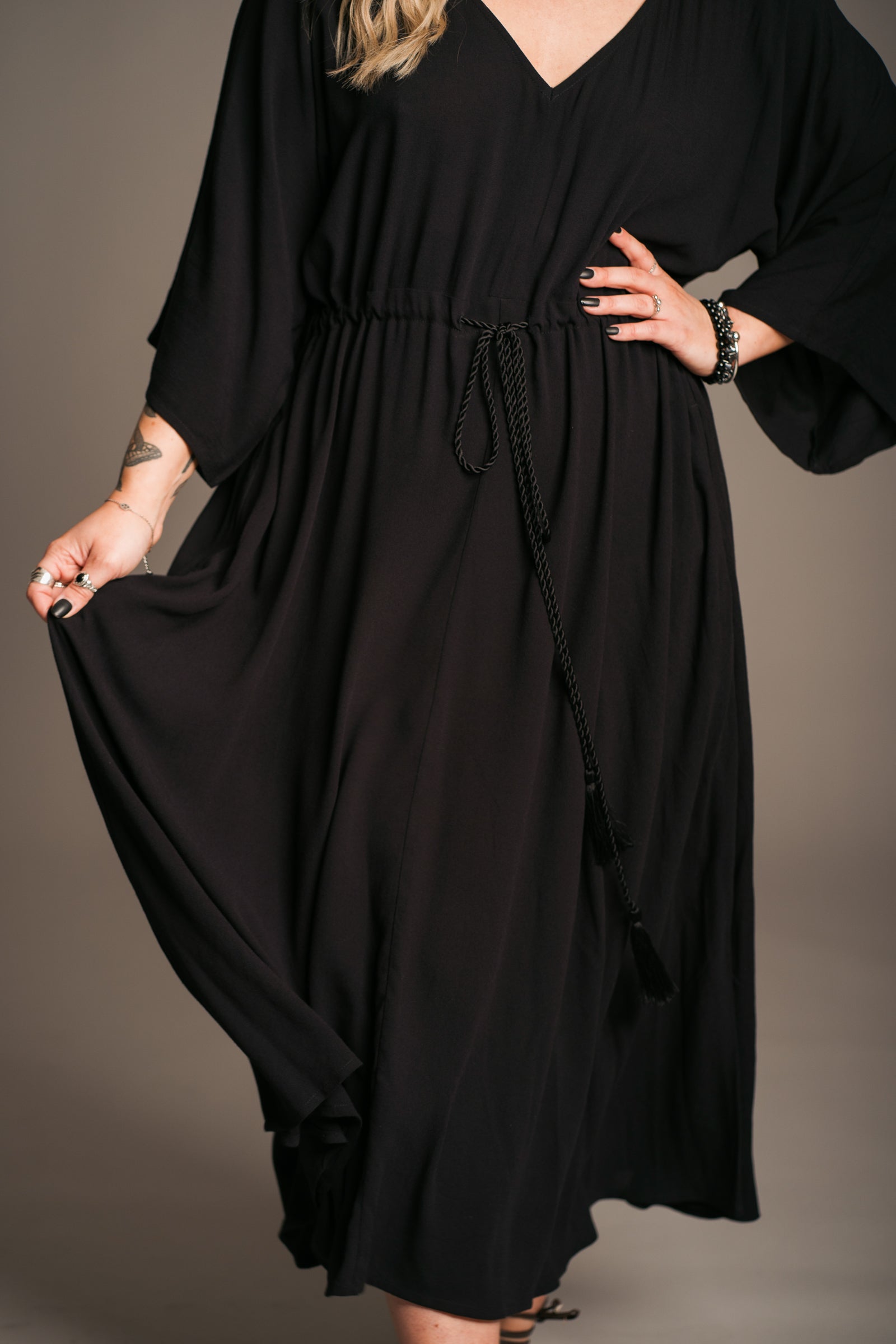 Model Erin wearing the Little Lies Blackbird Tie Waist Midi Dress, featuring a flattering V-neckline, batwing sleeves, an adjustable tie waist, and a flowing midi-length kaftan silhouette. This lightweight, drapey dress is styled with black ankle-tie sandals, exuding effortless bohemian elegance against a neutral backdrop.
