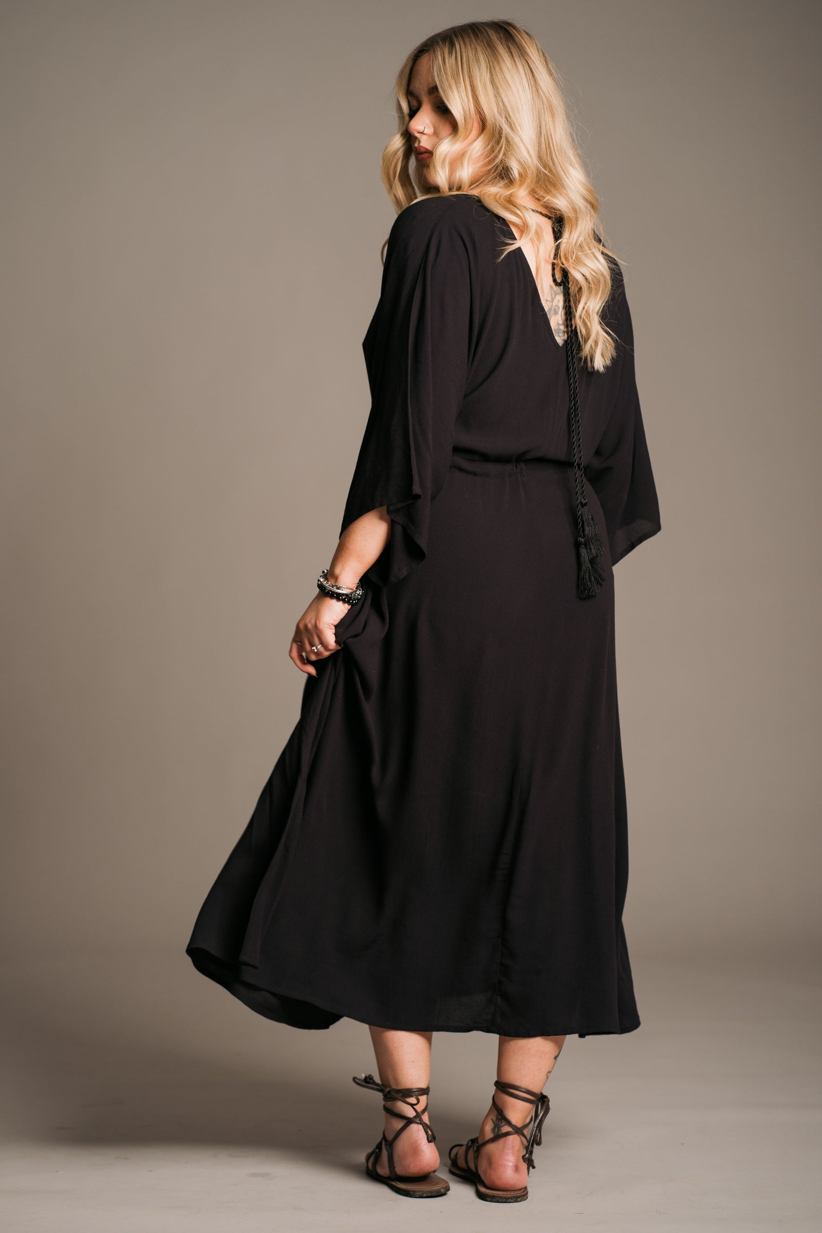 Model Erin wearing the Little Lies Blackbird Tie Waist Midi Dress showing the back of the dress, featuring a flattering V-neckline on the back a tie cord across the upper back, batwing sleeves, an adjustable tie waist, and a flowing midi-length kaftan silhouette. This lightweight, drapey dress is styled with black ankle-tie sandals, exuding effortless bohemian elegance against a neutral backdrop.