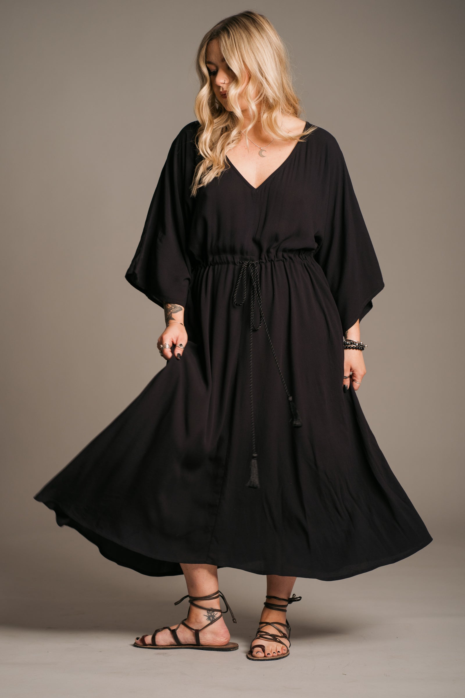 Model Erin wearing the Little Lies Blackbird Tie Waist Midi Dress, featuring a flattering V-neckline, batwing sleeves, an adjustable tie waist, and a flowing midi-length kaftan silhouette. This lightweight, drapey dress is styled with black ankle-tie sandals, exuding effortless bohemian elegance against a neutral backdrop.