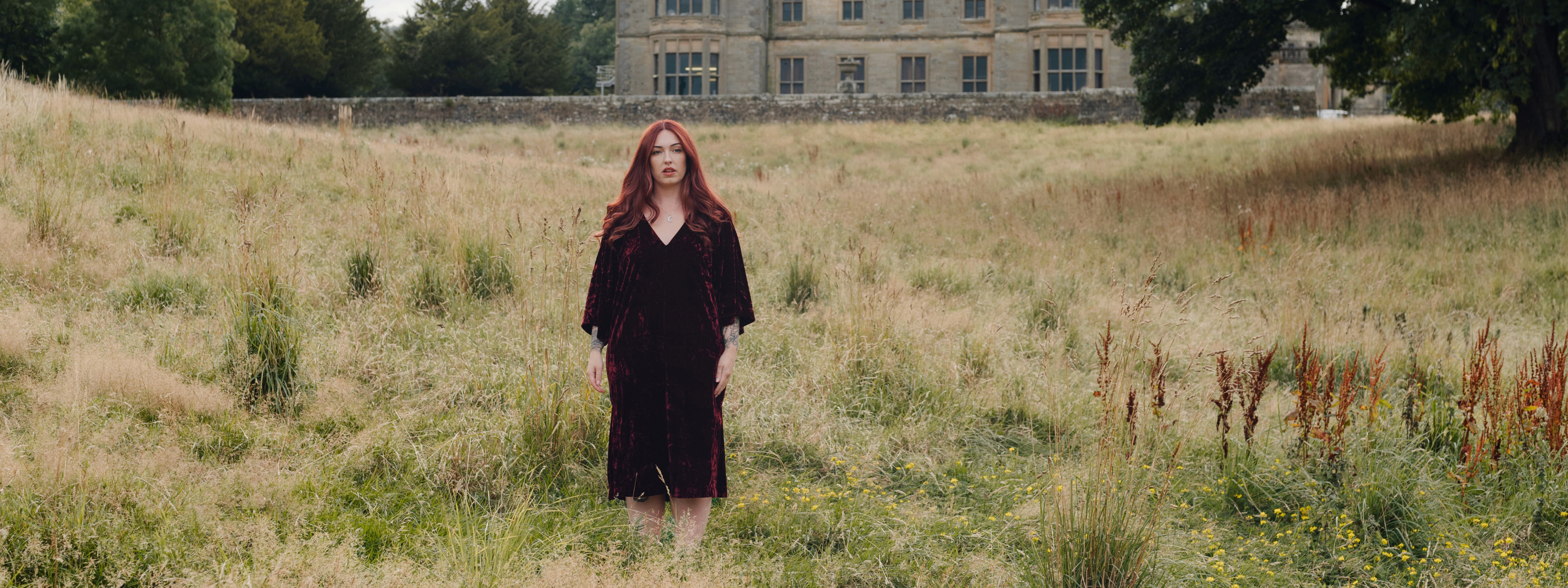 Taylor wears Little Lies Rumours Merlot Velvet Midi Dress styled outdoors in a meadow setting, showing the rich velvet texture and relaxed V‑neck silhouette