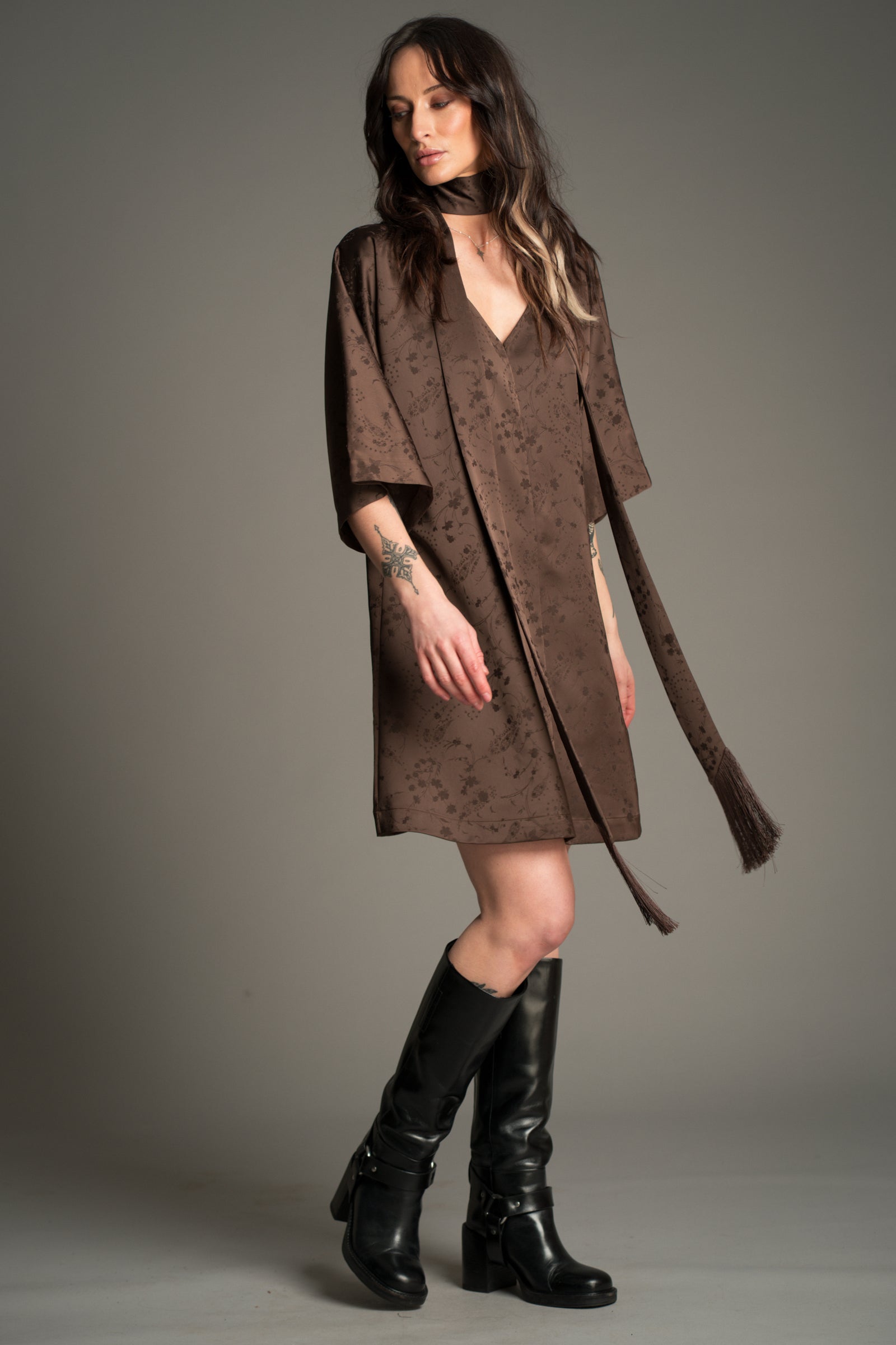 Victoria wears Little Lies Evermore Espresso Batwing Mini Dress featuring a relaxed V‑neckline, wide batwing sleeves, textured espresso‑brown fabric, and the tassel tie belt styled as a skinny scarf, with tall black boots.