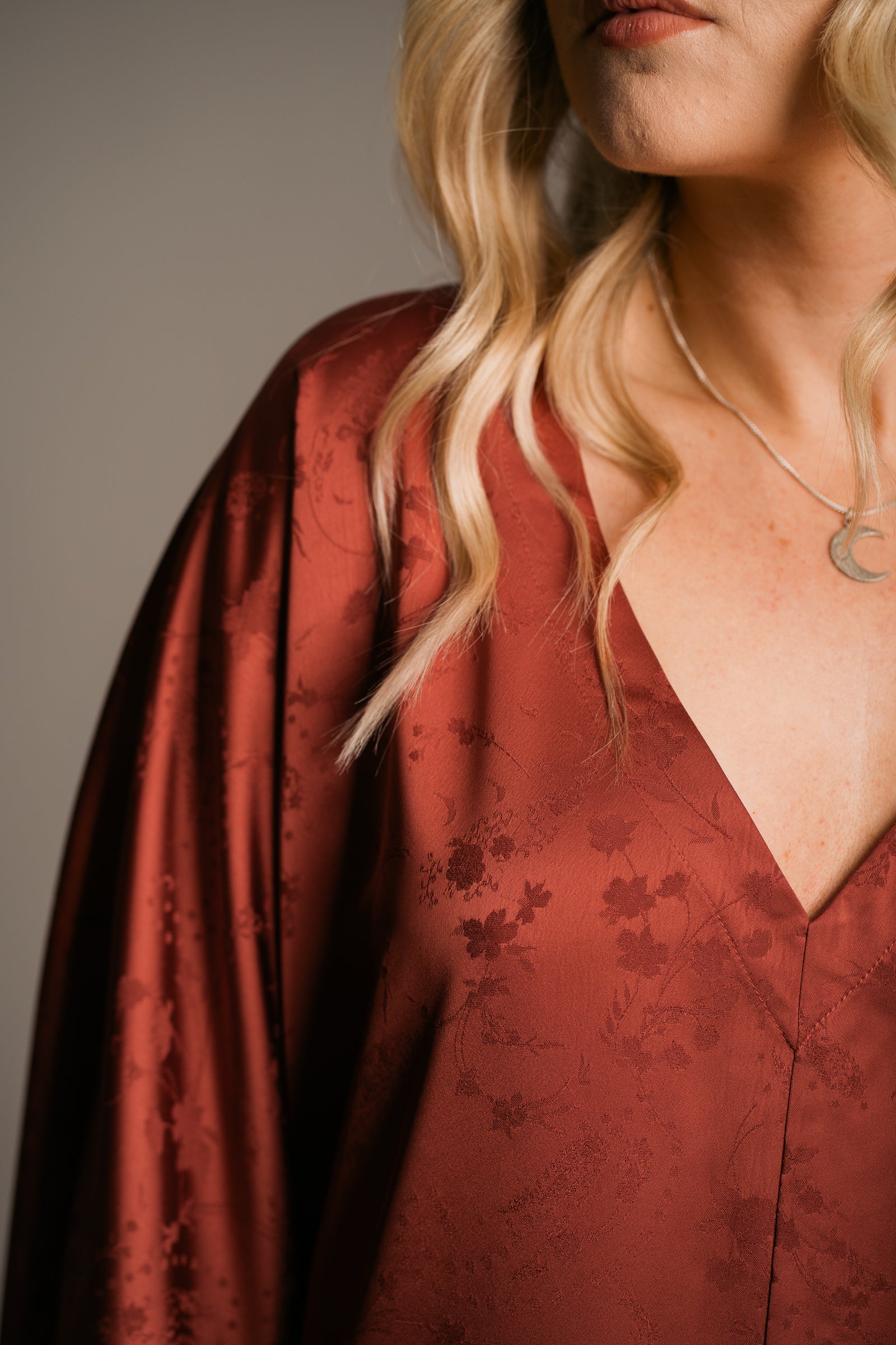 Our Model, Erin,  posing against a neutral backdrop, wearing the Little Lies Florence rust-colored fringe satin kimono dress this detail shot highlights the floral pattern in the fabric and the V-neckline. Paired with the Little Lies Crescent sterling silver necklace, the outfit is relaxed bohemian style. The model has long, wavy blonde hair.