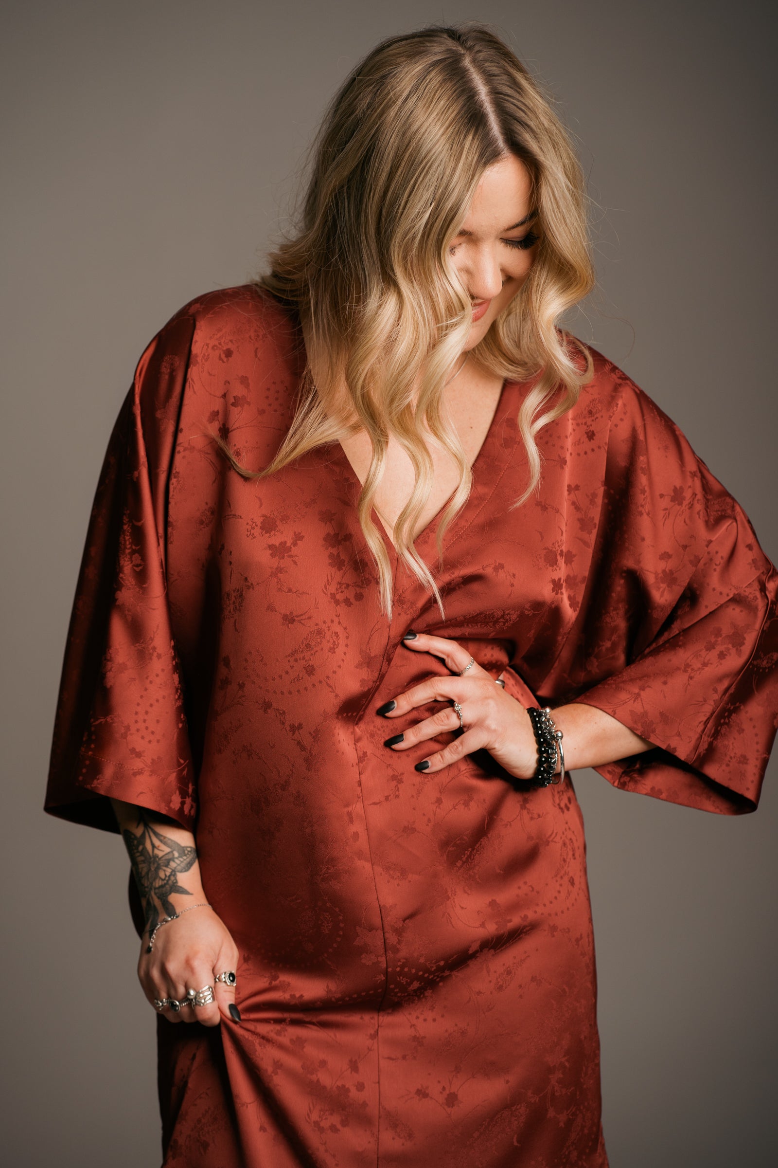 Our Model, Erin,  posing against a neutral backdrop, wearing the Little Lies Florence rust-colored fringe satin kimono dress with three-quarter sleeves and a V-neckline. Paired with  the Little Lies Crescent sterling silver necklace, relaxed bohemian style. The model has long, wavy blonde hair and a visible tattoos