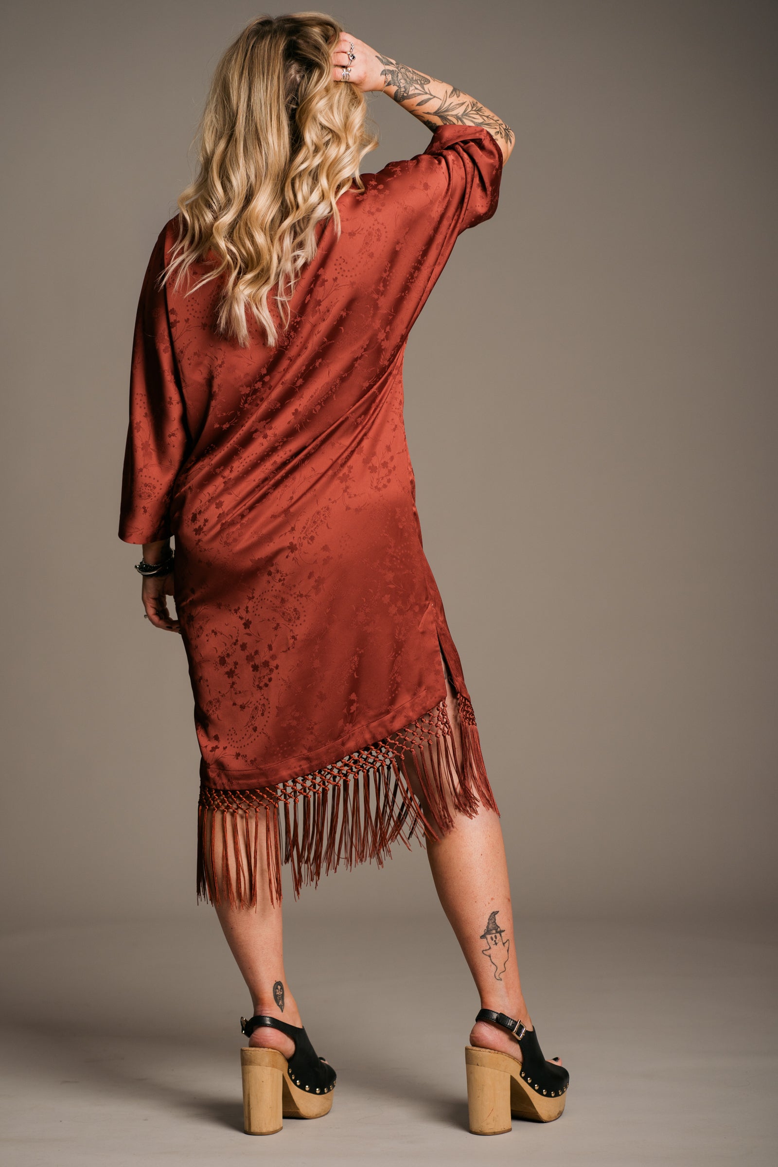 Our Model, Erin,  posing against a neutral backdrop, wearing the Little Lies Florence rust-colored fringe satin kimono dress with three-quarter sleeves and fringing along the hem. Paired with black wooden platform clogs, the outfit features a relaxed bohemian style. The model has long, wavy blonde hair and a visible tattoos.