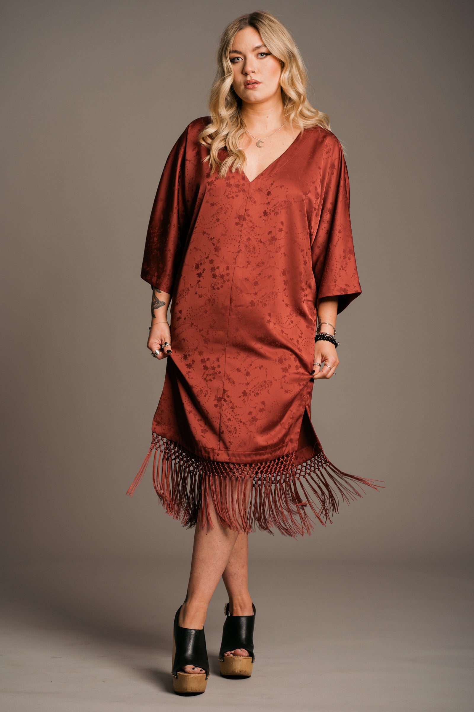 Our Model, Erin,  posing against a neutral backdrop, wearing the Little Lies Florence rust-colored fringe satin kimono dress with three-quarter sleeves and a V-neckline. Paired with black wooden platform clogs, and the Little Lies Crescent sterling silver necklace, perfect relaxed bohemian style. The model has long, wavy blonde hair and a visible tattoos on the forearm.