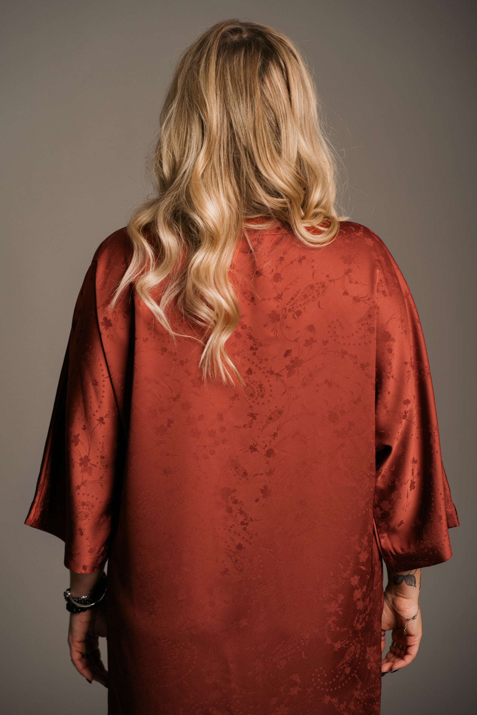Our Model, Erin,  posing against a neutral backdrop, wearing the Little Lies Florence rust-colored fringe satin kimono dress with three-quarter sleeves. The model has long, wavy blonde hair, stacks of jewellery and visible tattoos.