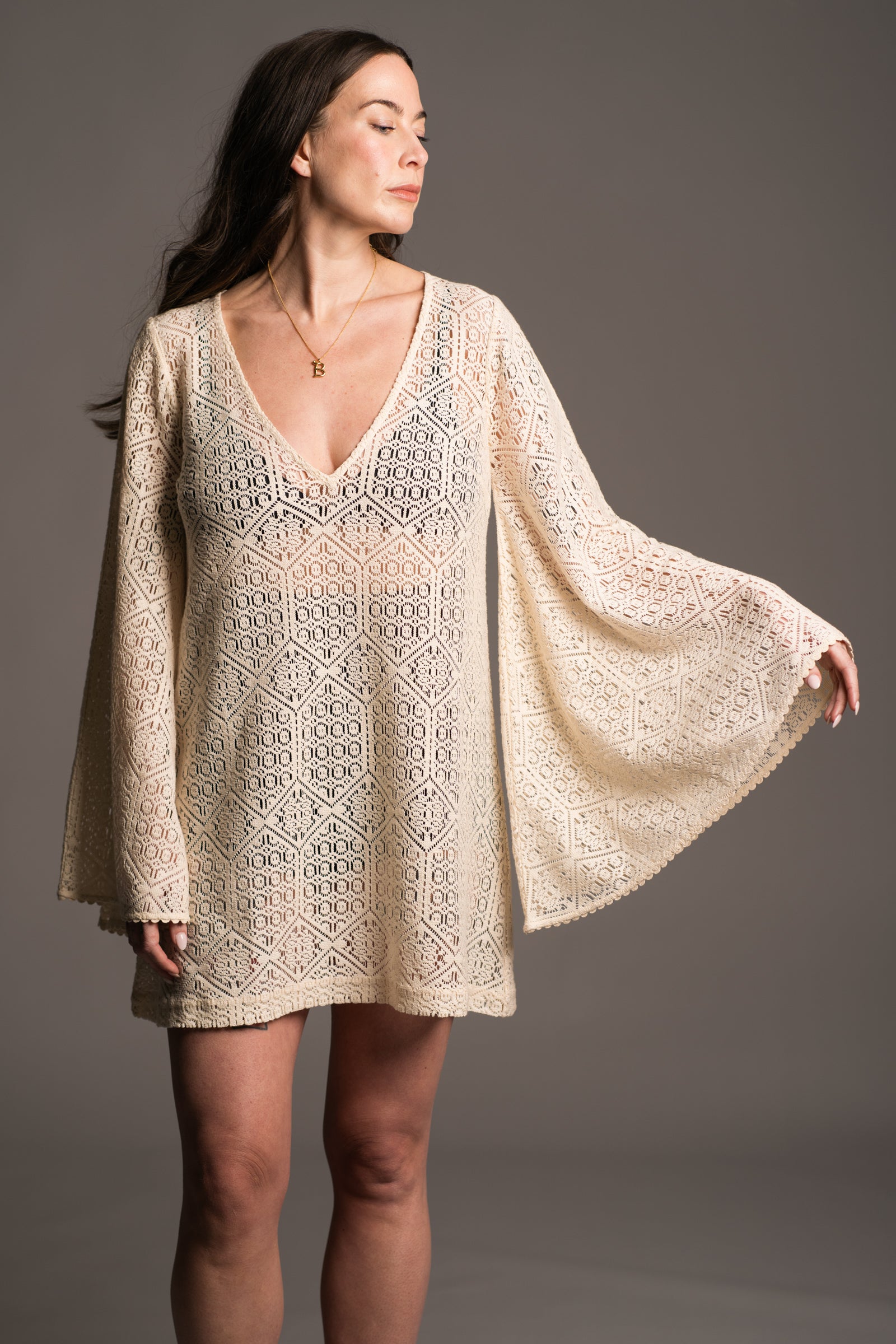 Our model, Bex, standing against a neutral backdrop in the studio wearing the Little Lies Goldie Ivory Crochet Bell Sleeved Mini Dress, featuring intricate crochet detailing, flowing bell sleeves, a V-neckline and a flattering mini silhouette. Showing the extreme bell sleeve detail and styled with gold 'B' necklace the look embodies effortless bohemian elegance.