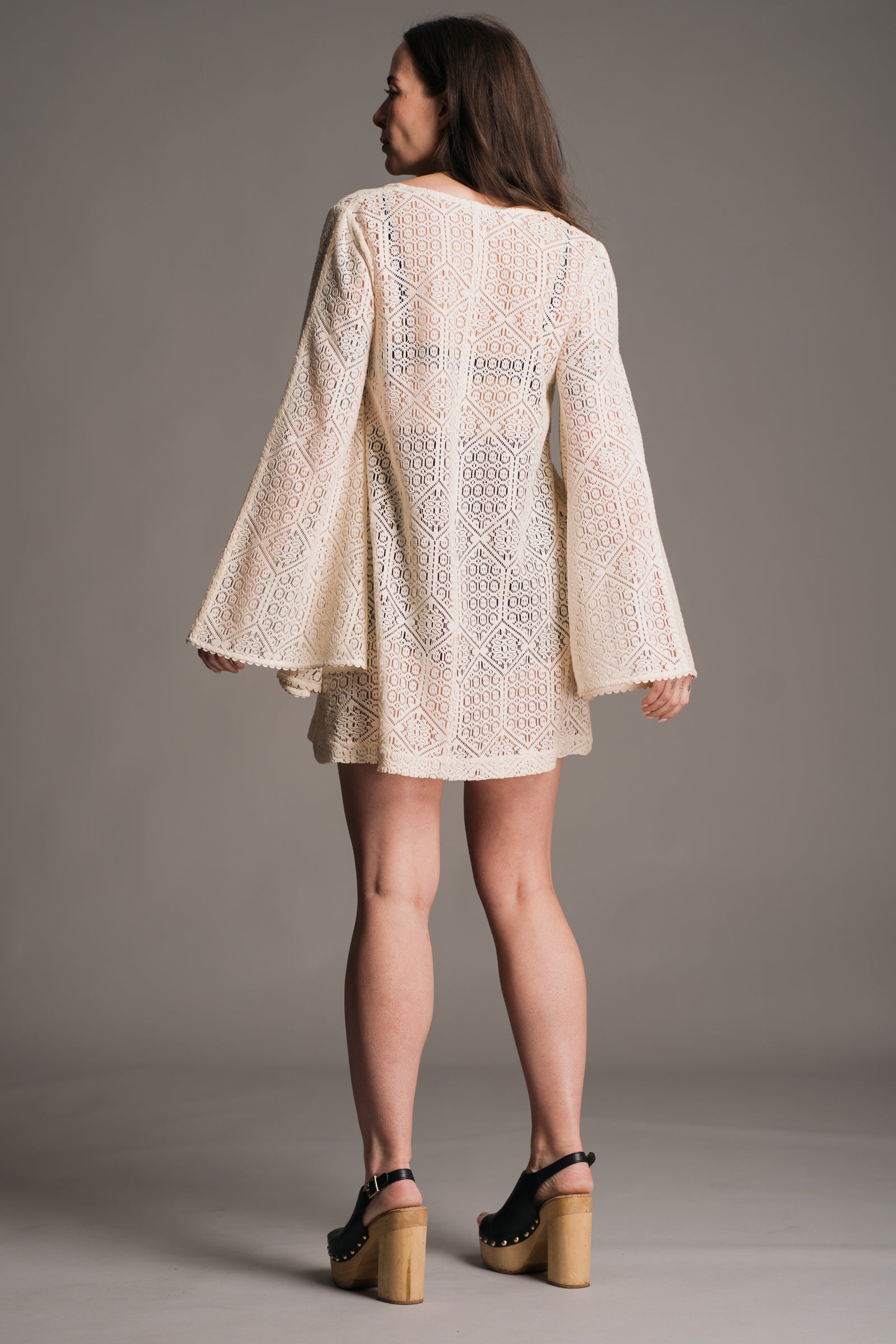 Our model, Bex, standing against a neutral backdrop in the studio showing the back of the Little Lies Goldie Ivory Crochet Bell Sleeved Mini Dress, featuring intricate crochet detailing, flowing bell sleeves, and a flattering mini silhouette. Styled with vintage black wooden clogs the look embodies effortless bohemian elegance.