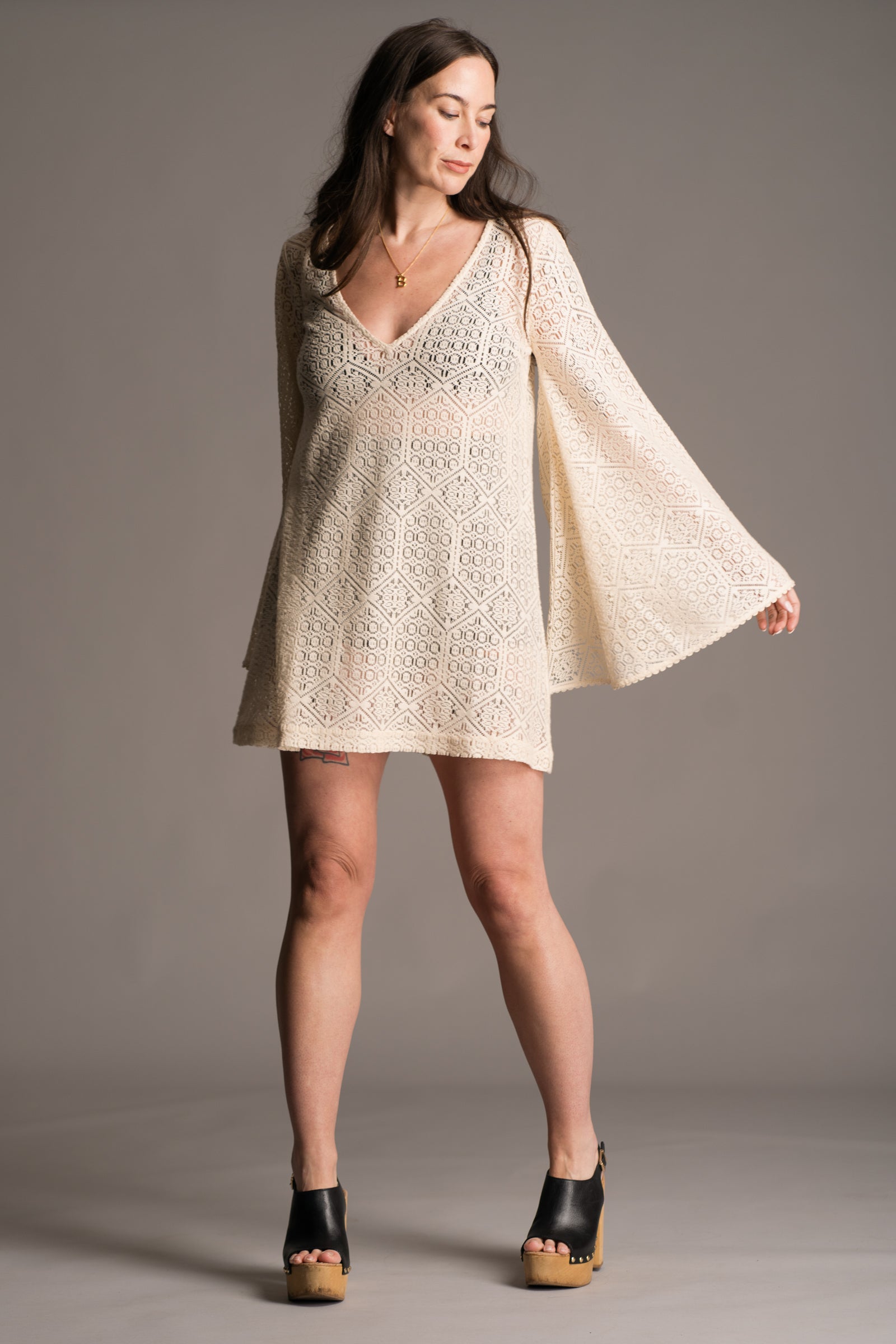 Our model, Bex, standing against a neutral backdrop in the studio wearing the Little Lies Goldie Ivory Crochet Bell Sleeved Mini Dress, featuring intricate crochet detailing, flowing bell sleeves, a V-neckline and a flattering mini silhouette. Styled with vintage black wooden clogs the look embodies effortless bohemian elegance.
